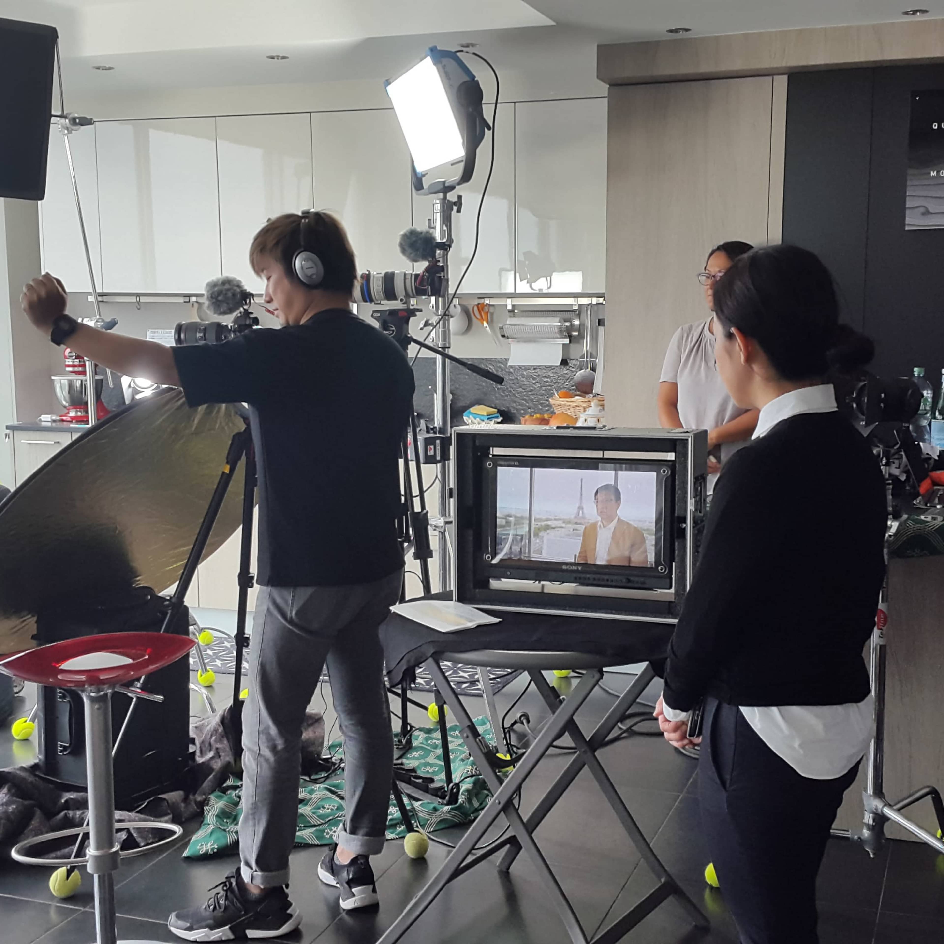 A film crew sets up a shoot in a modern kitchen, with a camera pointed at a seated subject and various lighting equipment positioned around.
