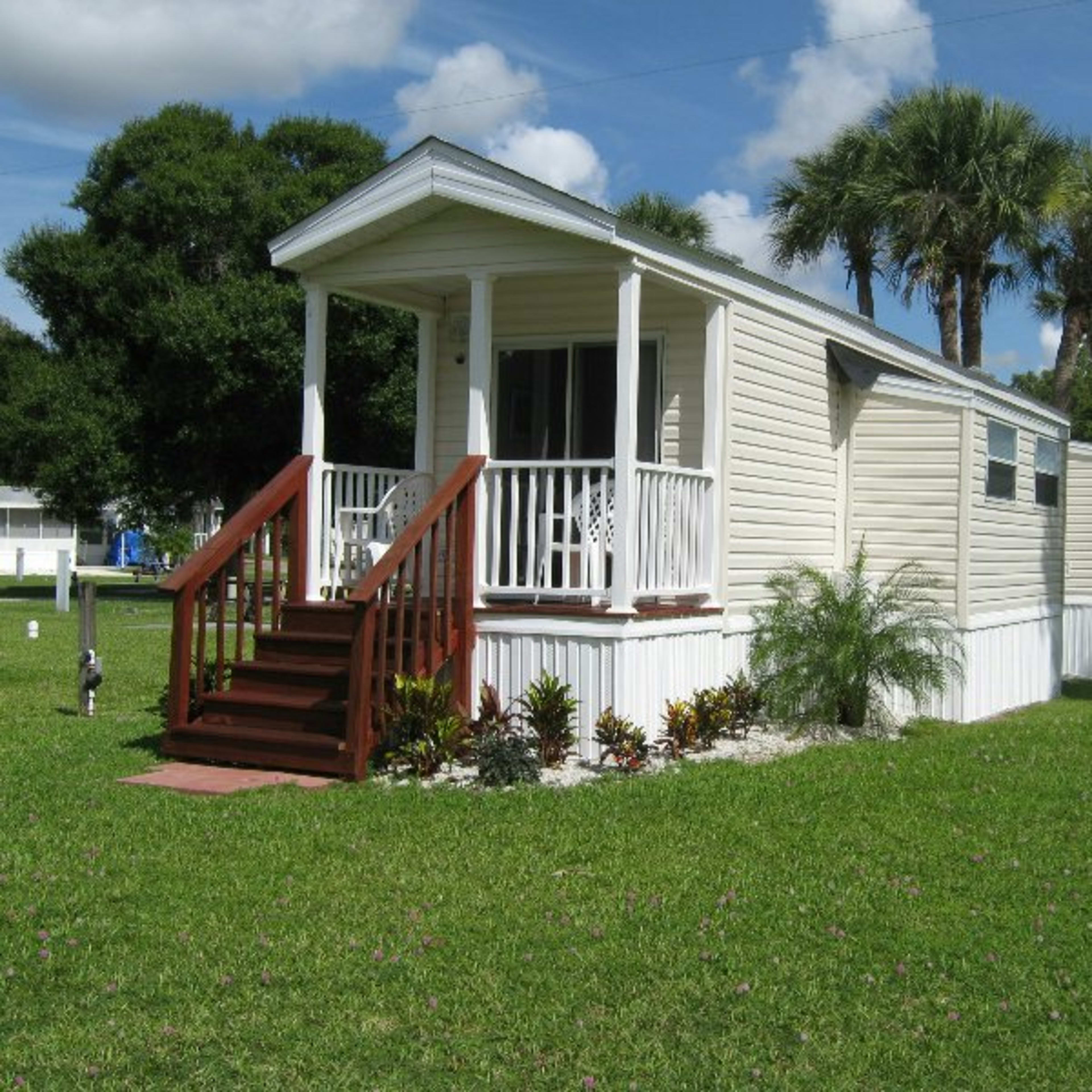 A small white manufactured home with a front porch and wooden steps is set on a grassy lawn surrounded by palm trees.