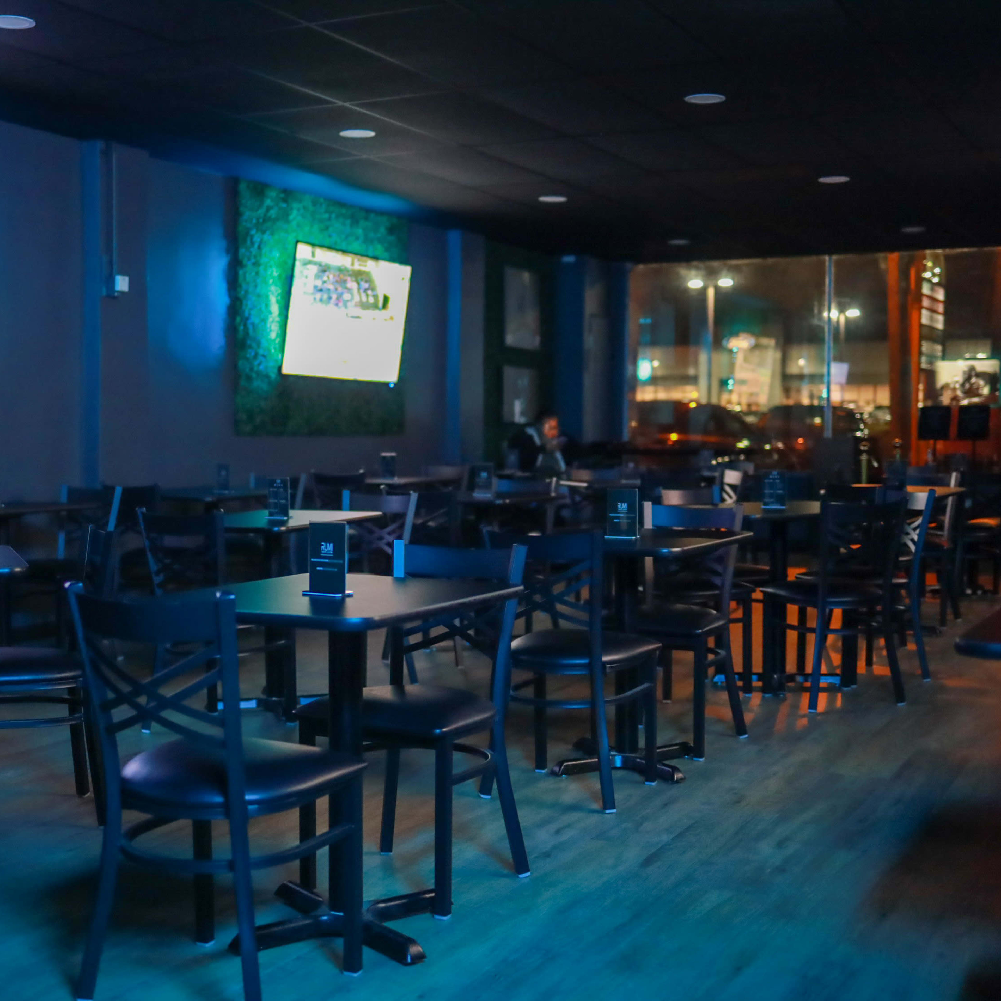 An empty bar with black tables and chairs, featuring two televisions on the walls and large windows displaying a city view at night.