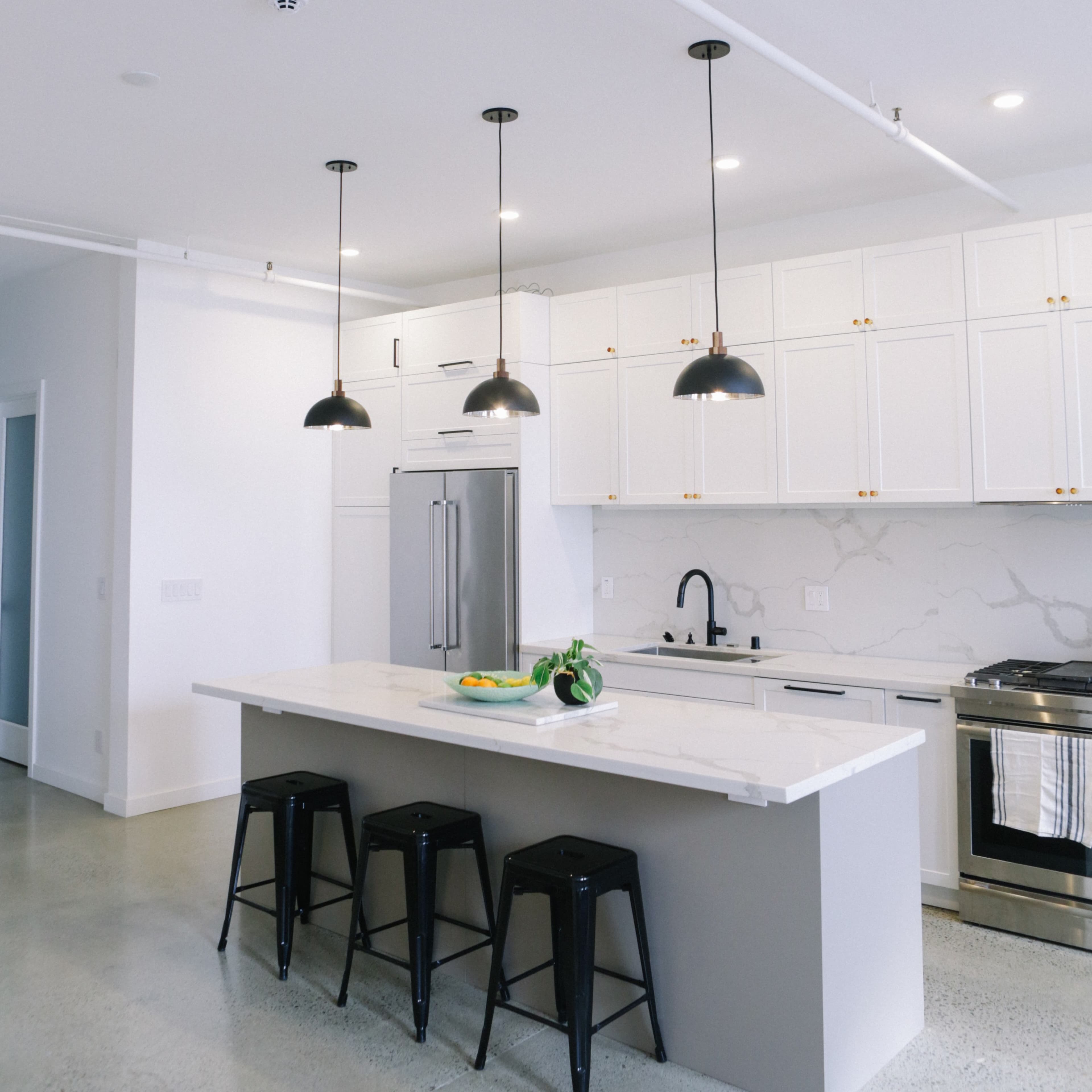 A modern kitchen features a large island with a white countertop, black pendant lights overhead, and minimalistic black stools.