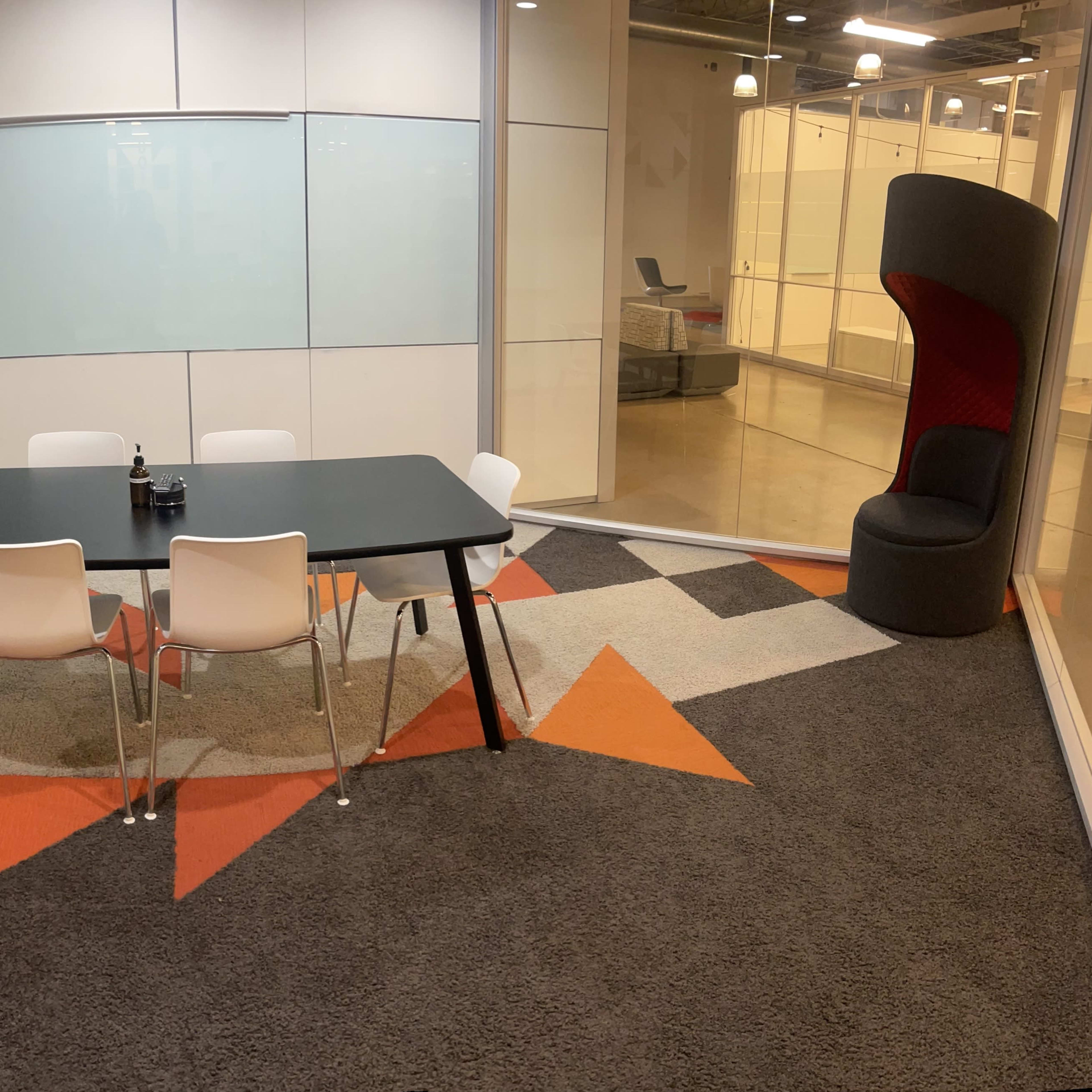 A modern meeting room with a black table surrounded by white chairs on a patterned orange and gray carpet, featuring a wall-mounted TV and a separate red acoustic chair in one corner.
