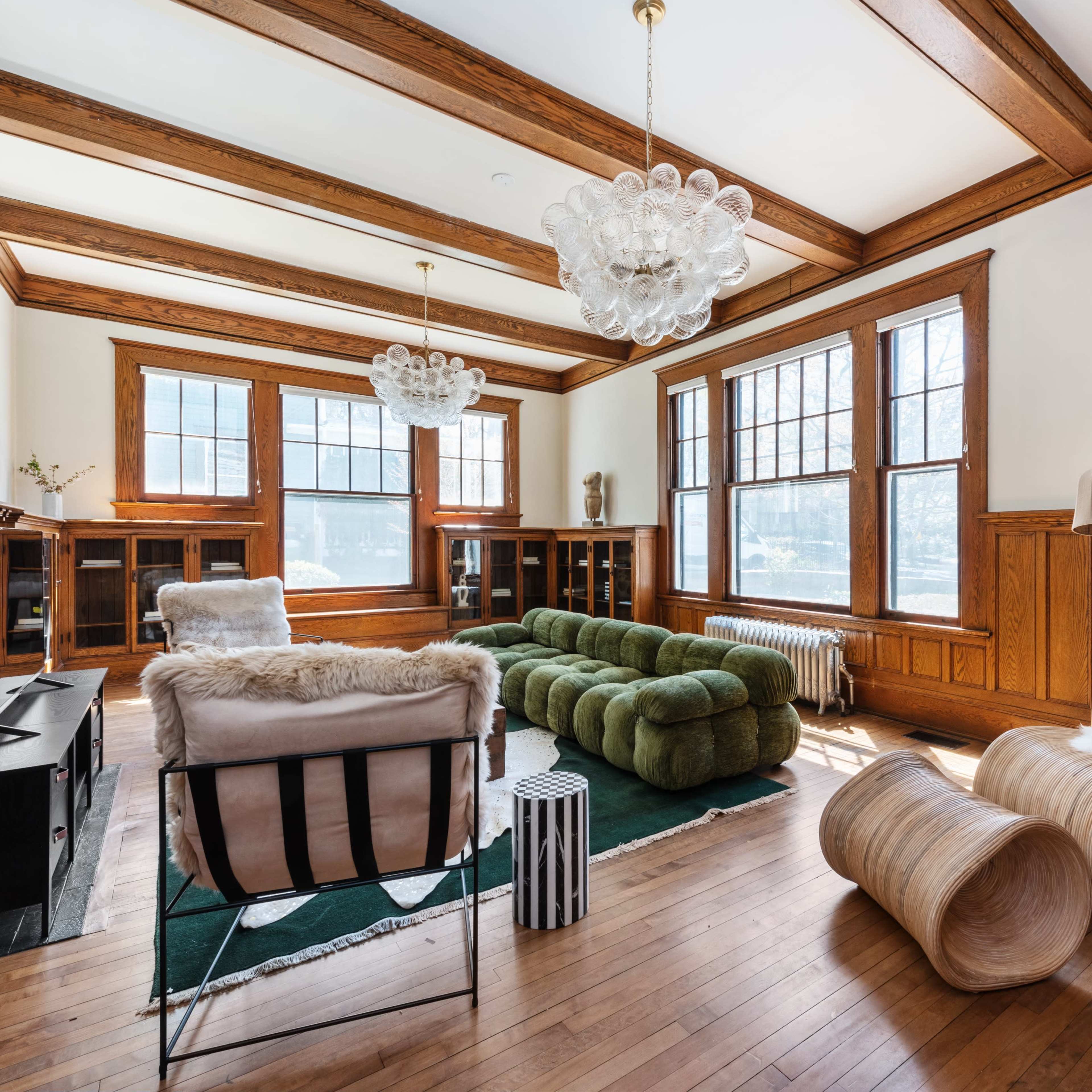 A spacious living room featuring wooden beams, large windows, a green sectional sofa, and various modern furniture pieces.