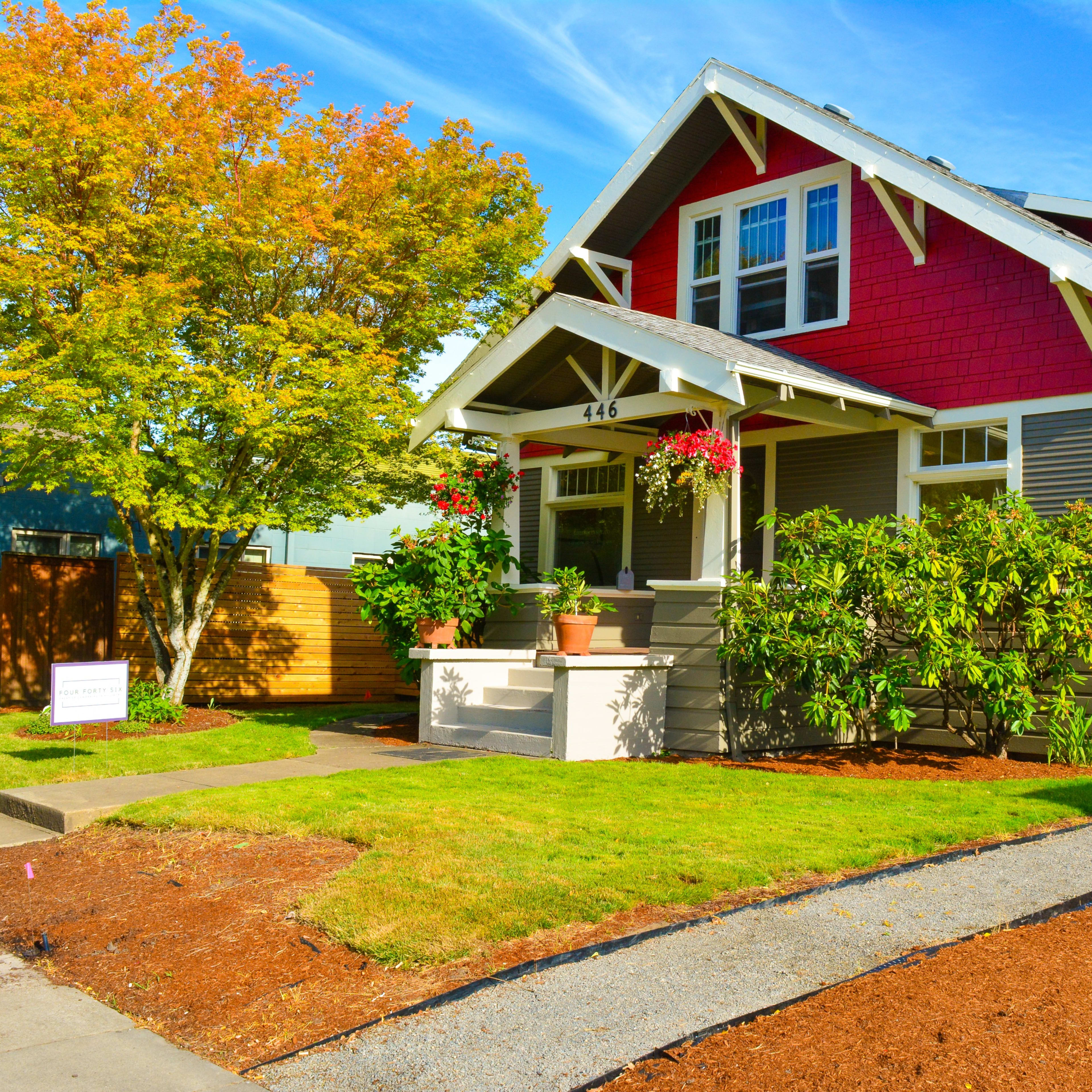 A house with a red and gray exterior, surrounded by green plants and trees, sits on a well-maintained lawn.