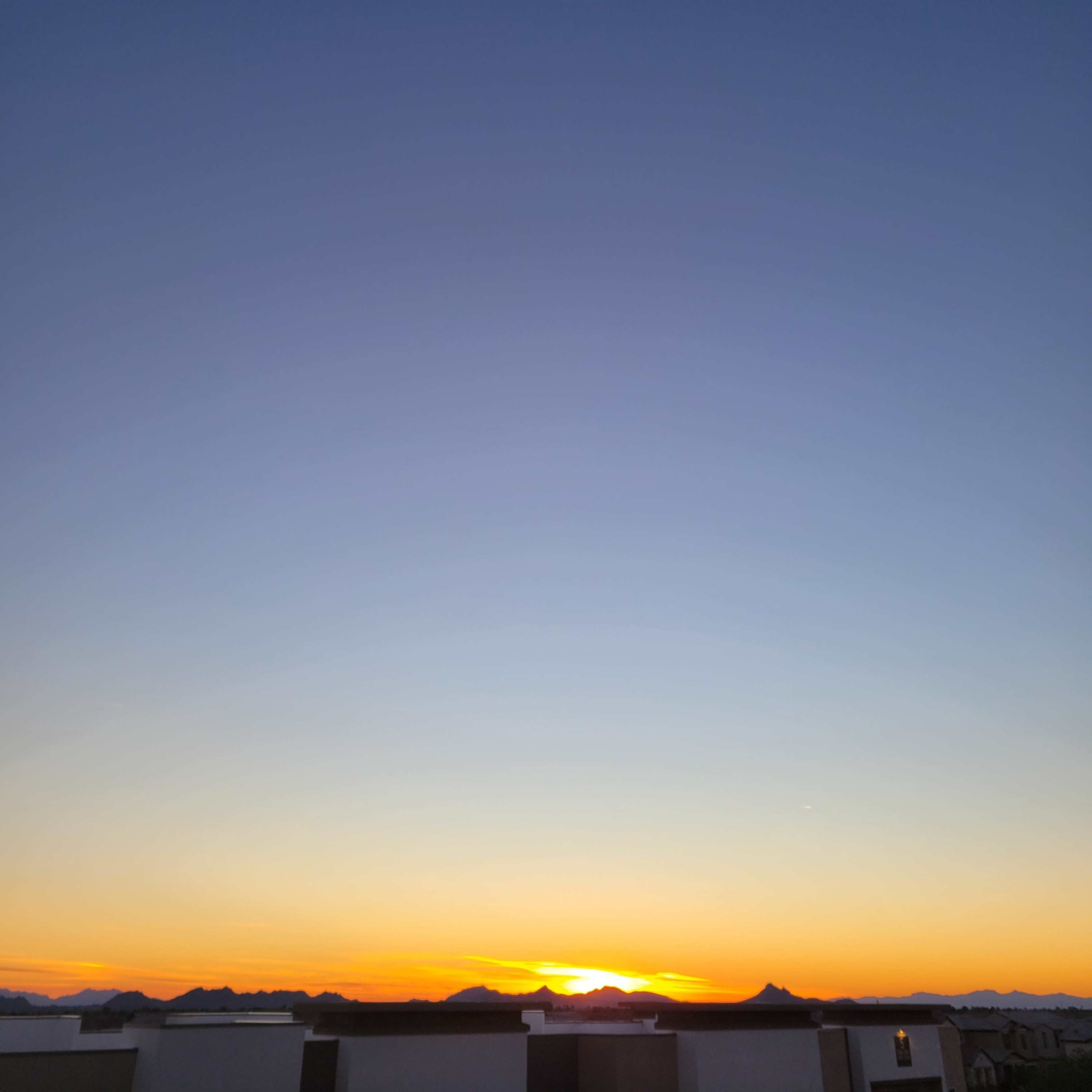 A sunset over a mountainous horizon with a clear sky transitioning from blue to orange.