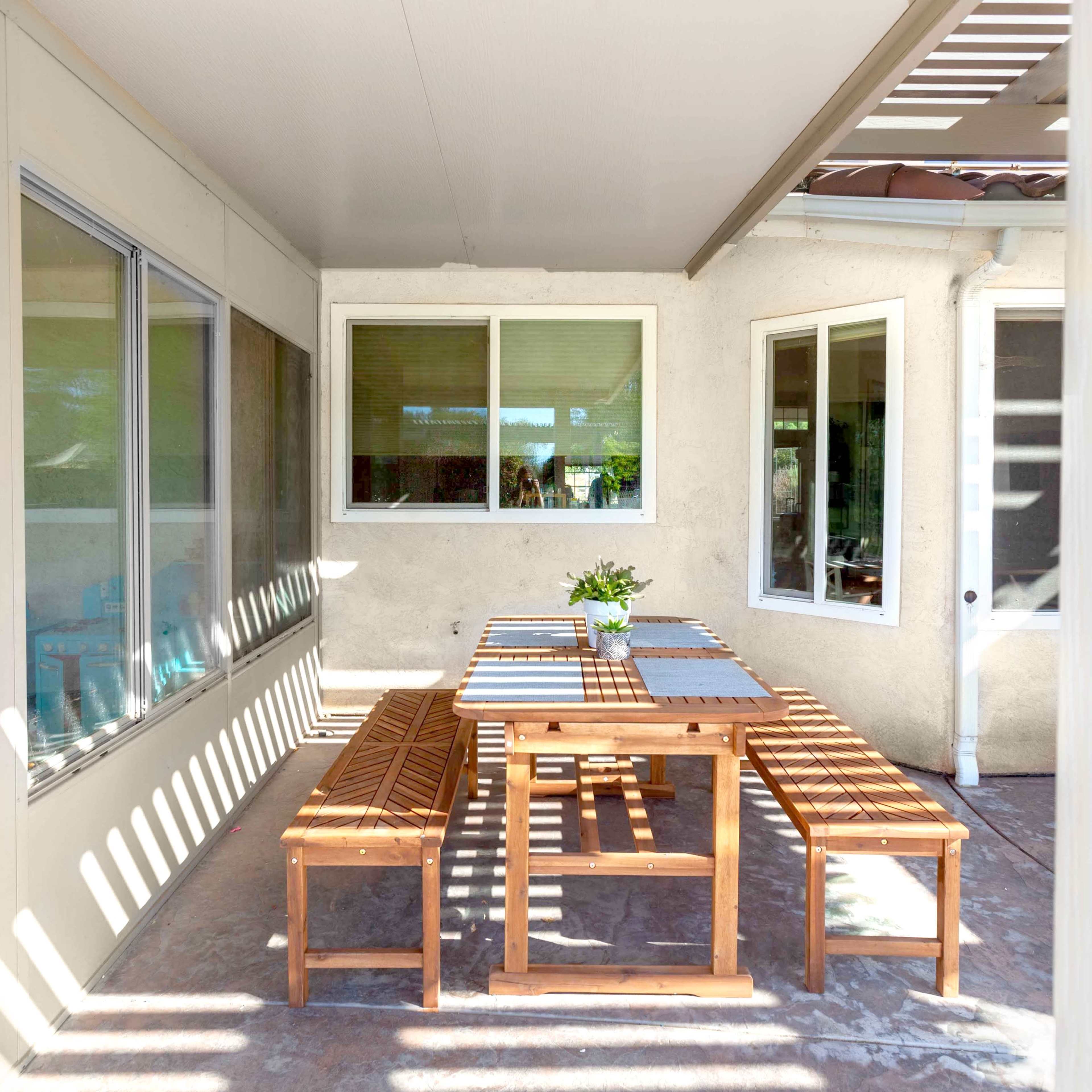 A wooden dining table is set with benches in a covered patio area, surrounded by large windows and natural light.