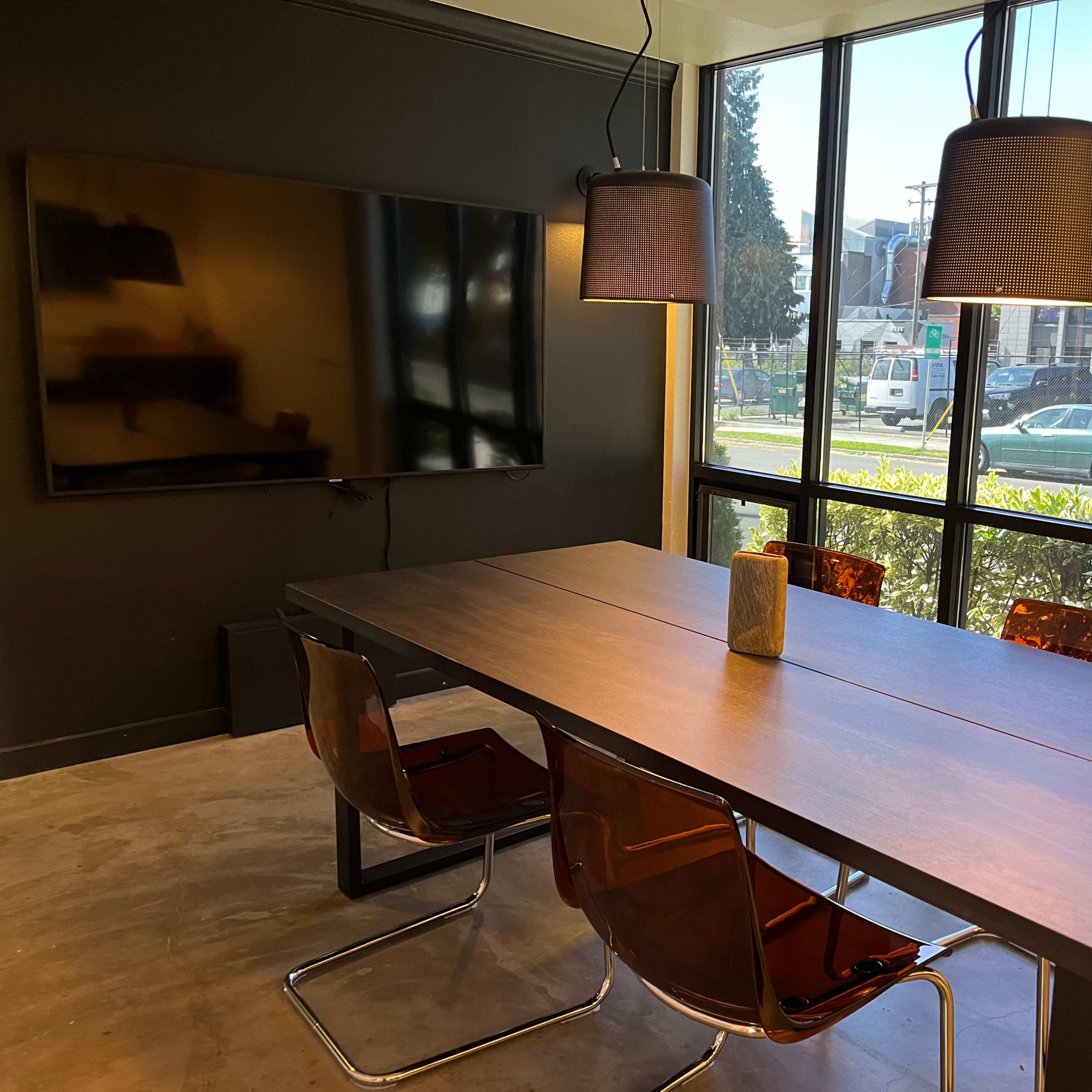 A modern conference room features a long table with four chairs, a television mounted on the wall, and large windows overlooking an outdoor area.