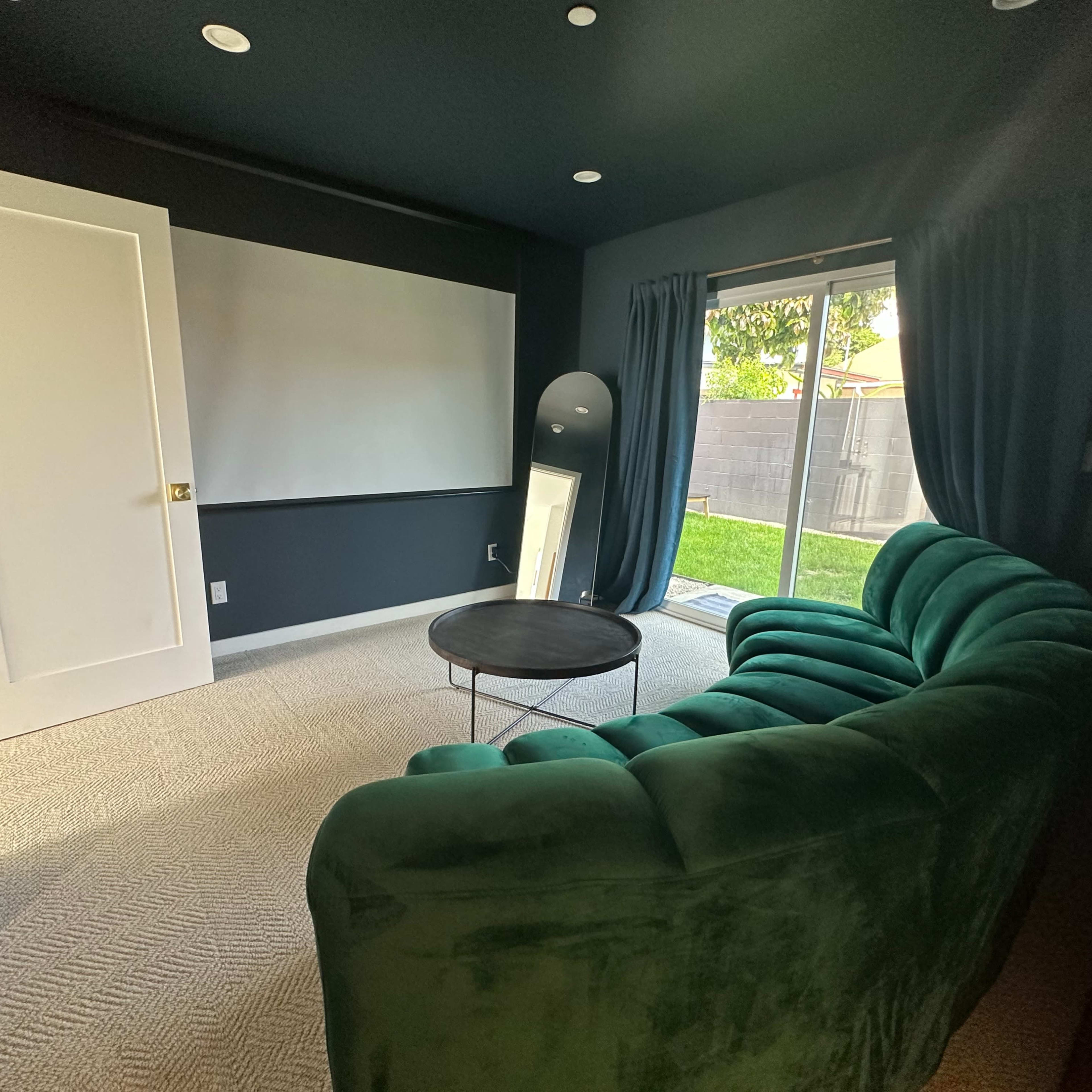 The room features a green velvet sofa, a round coffee table, a blank wall for projection, and large windows with curtains looking out to a grassy area.