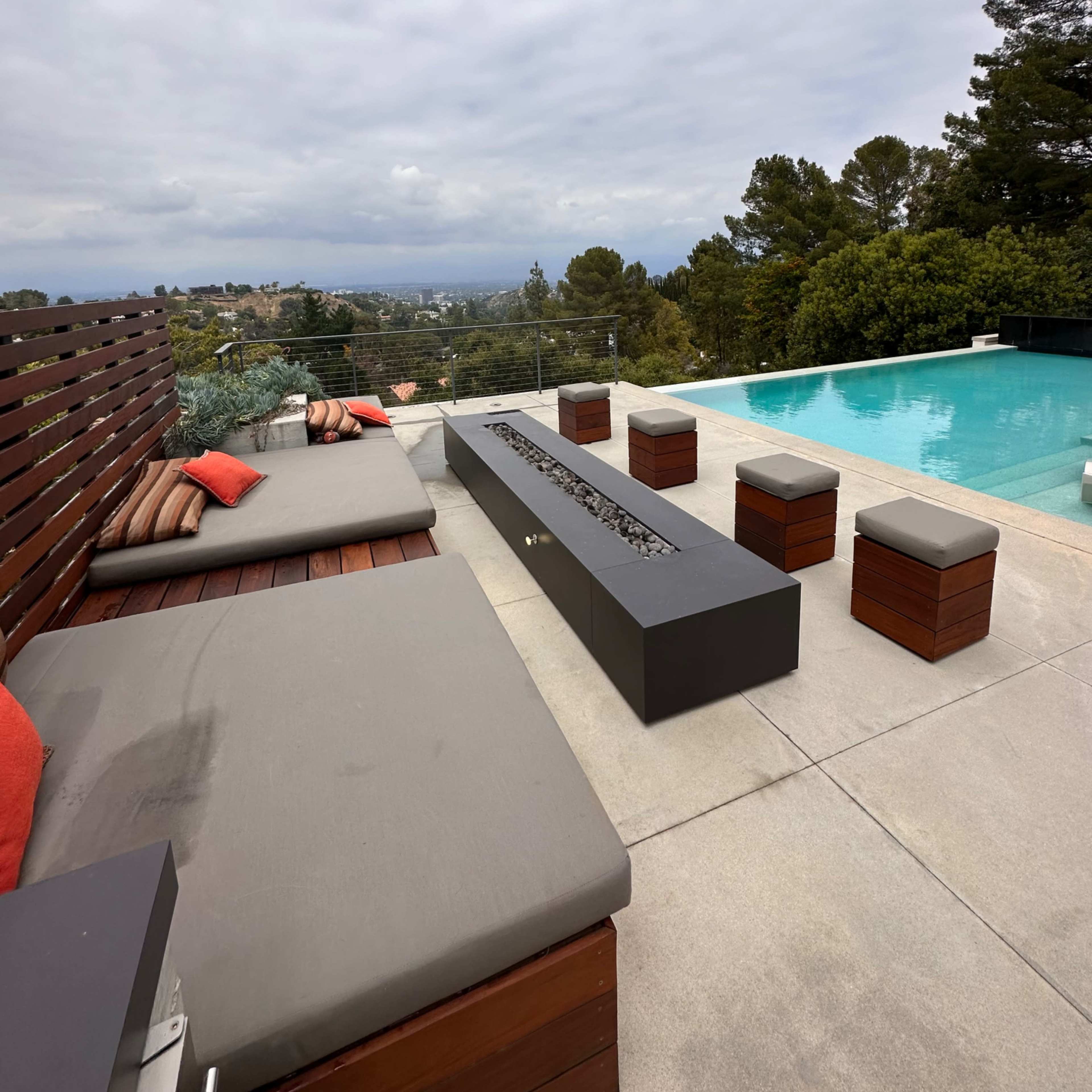 A modern outdoor patio features a lounge area with cushions and a fire pit, overlooking a pool and scenic landscape.