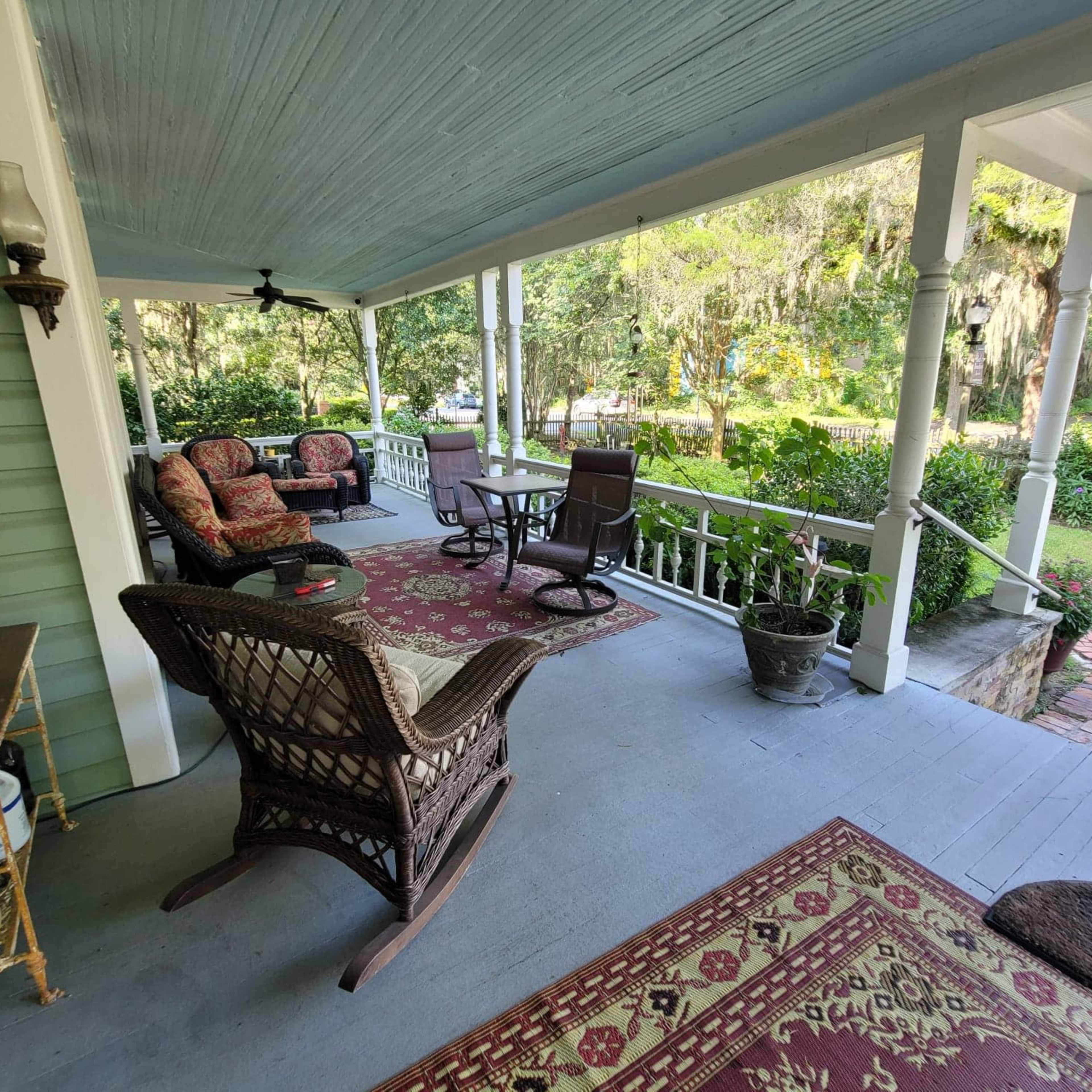 A covered porch with seating arrangements, including wicker chairs and cushions, overlooking a garden.