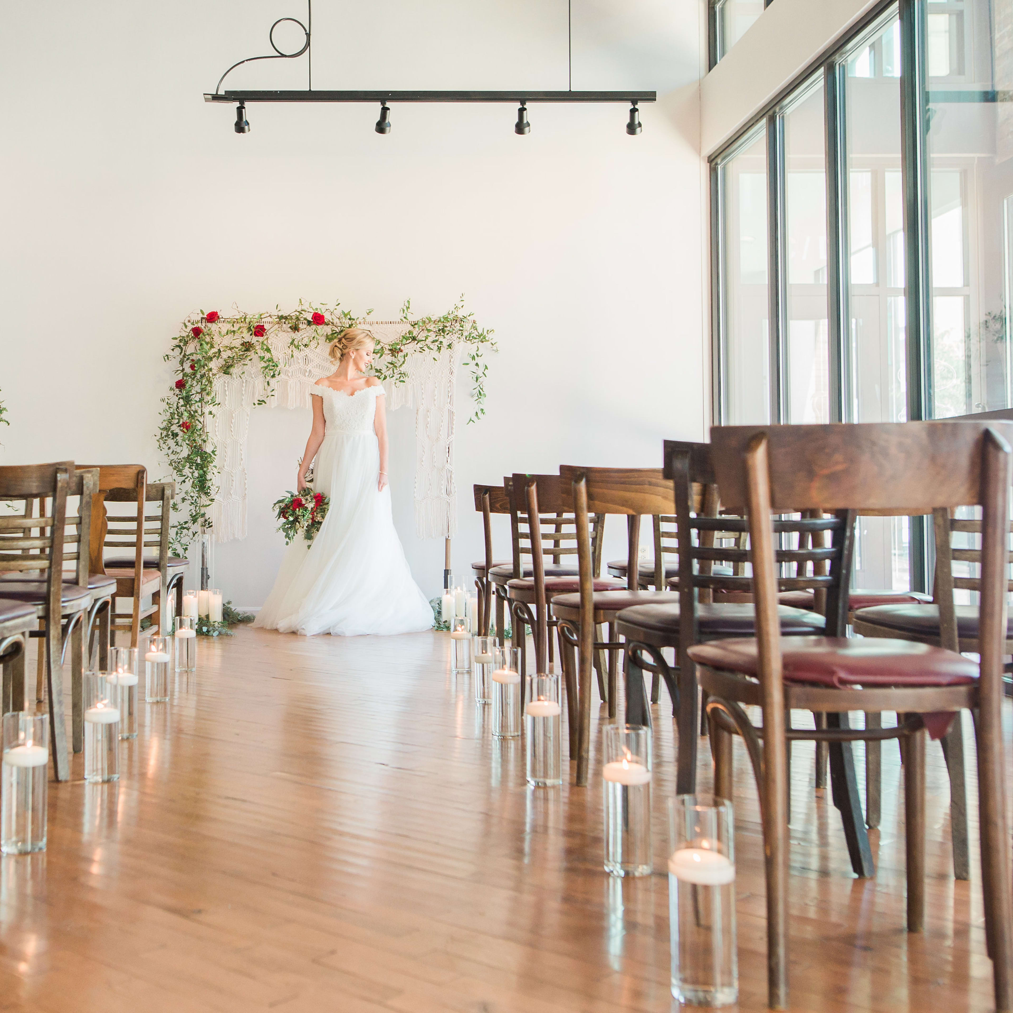 A bride stands in front of a floral archway at the end of an aisle lined with candles in a spacious, well-lit venue.