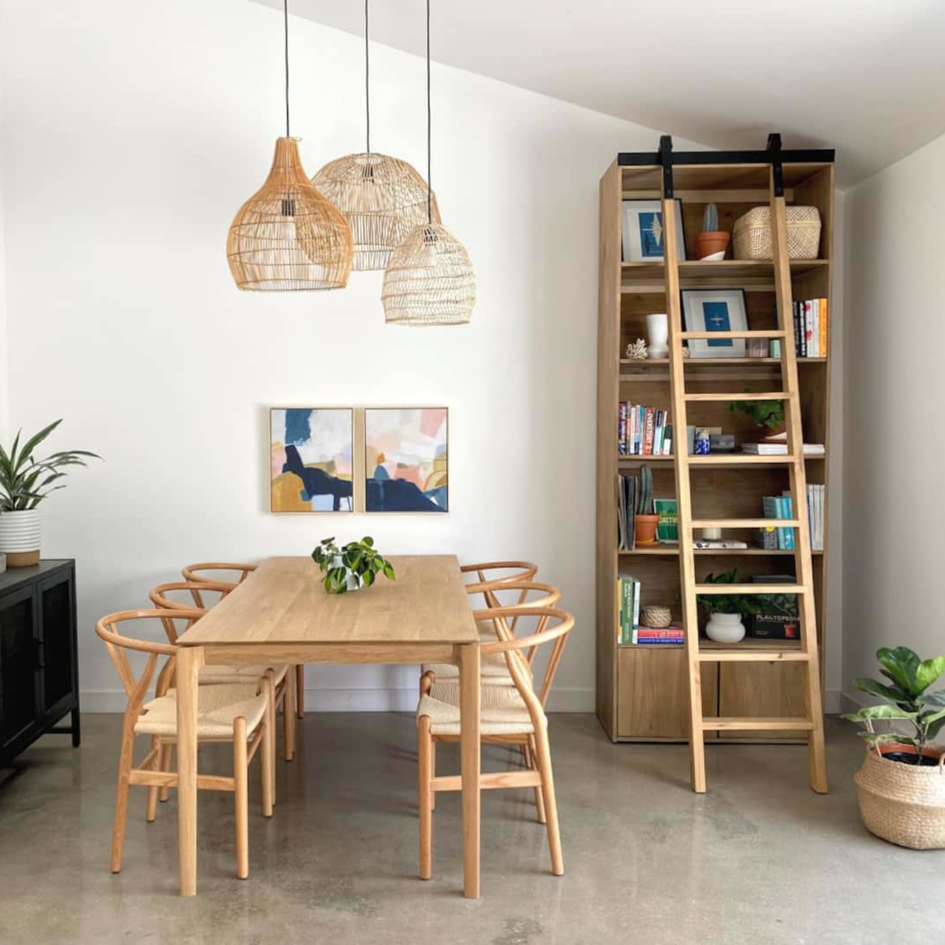 A modern dining area features a wooden table surrounded by six chairs, a tall bookshelf filled with books, and pendant lights hanging from the ceiling.