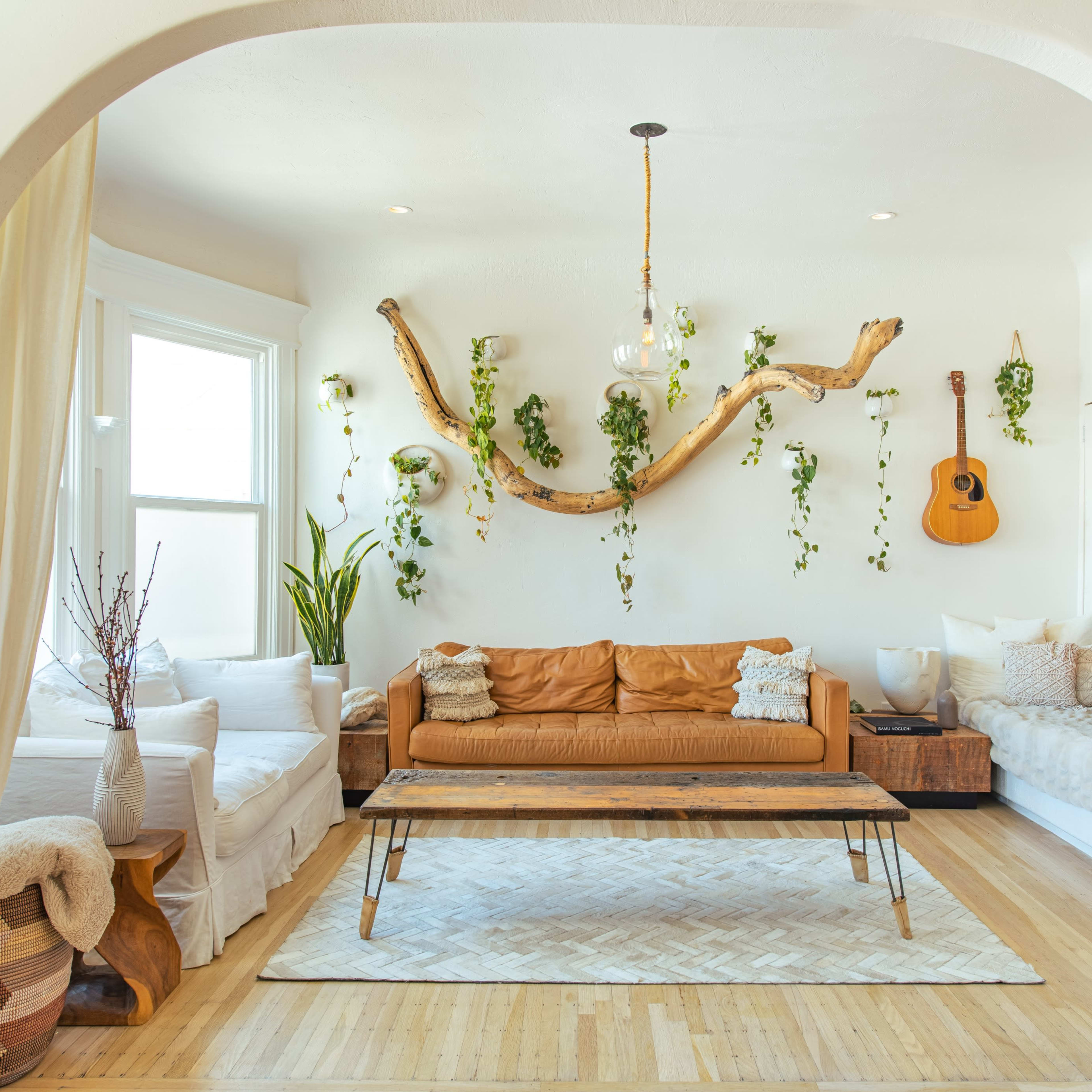 A bright living room features a leather sofa, a wooden coffee table, and plants hanging on the walls, with a guitar and a fireplace as focal points.