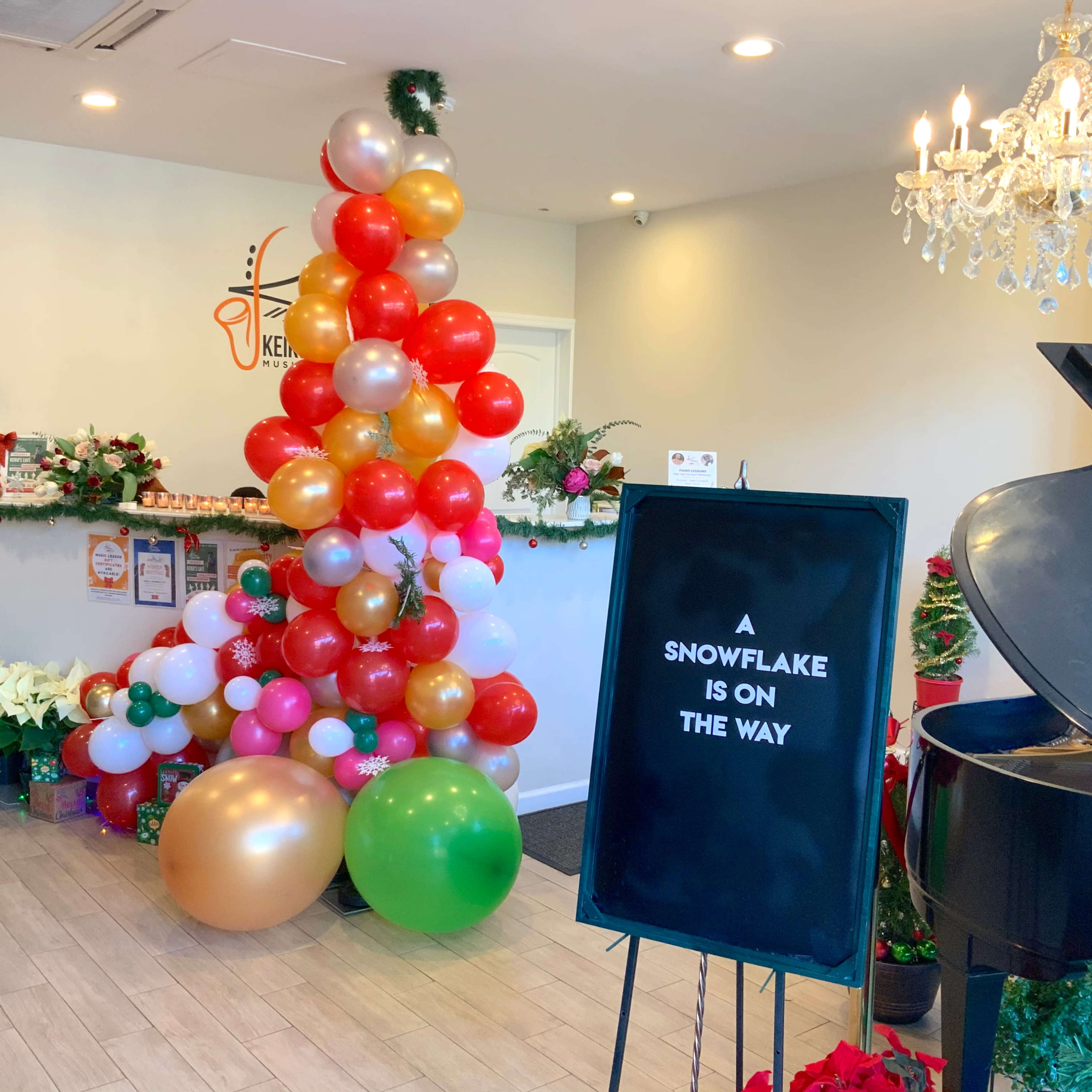 A festive interior of a café decorated with a balloon arch and a sign that reads "A SNOWFLAKE IS ON THE WAY" near a piano.