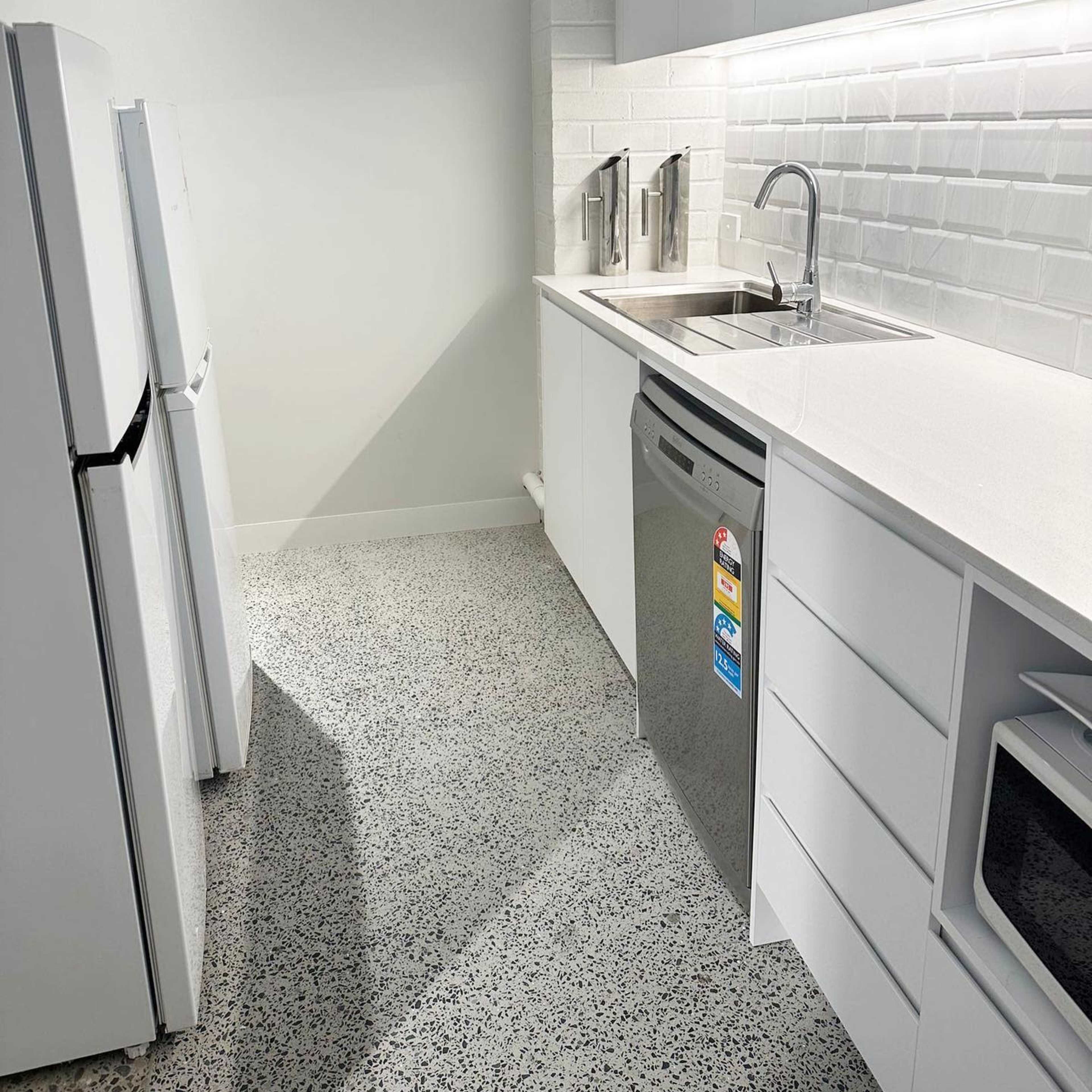 A compact kitchen features white cabinets, a stainless steel dishwasher, and a refrigerator, with a light-colored countertop and backsplash.