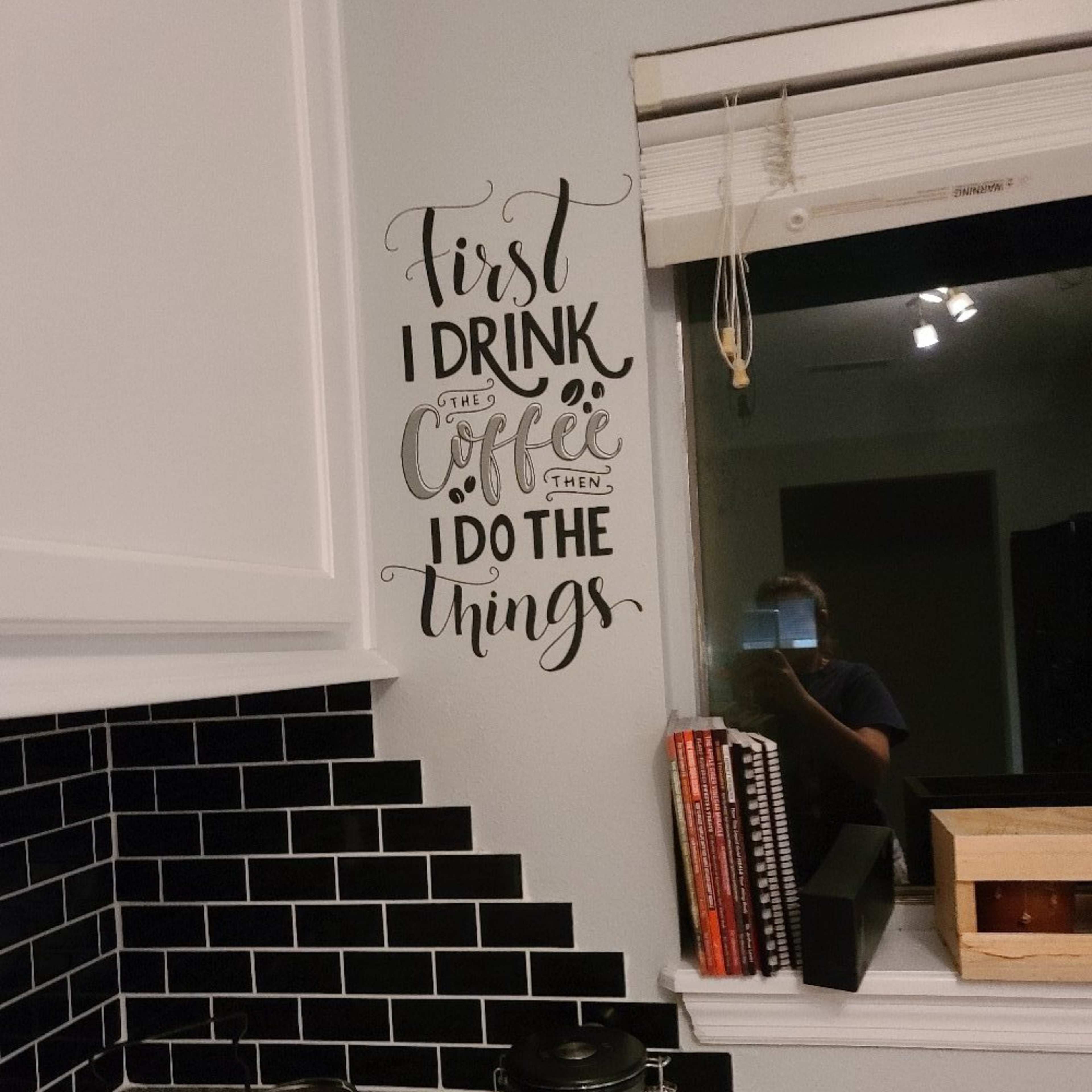 A kitchen corner features a wall decal that reads, "First I drink the coffee, then I do the things," beside a window and shelf with various items.