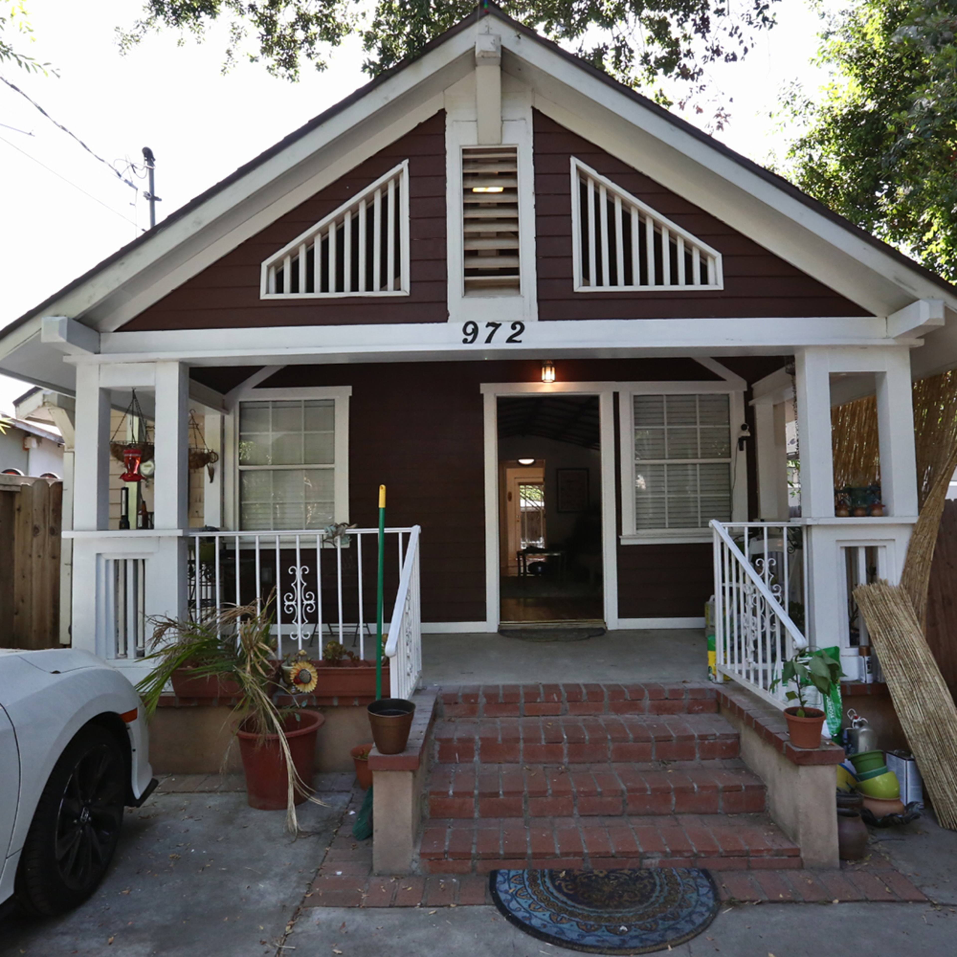 A single-story brown house with a white porch and steps, featuring a front door and several plants on the porch.