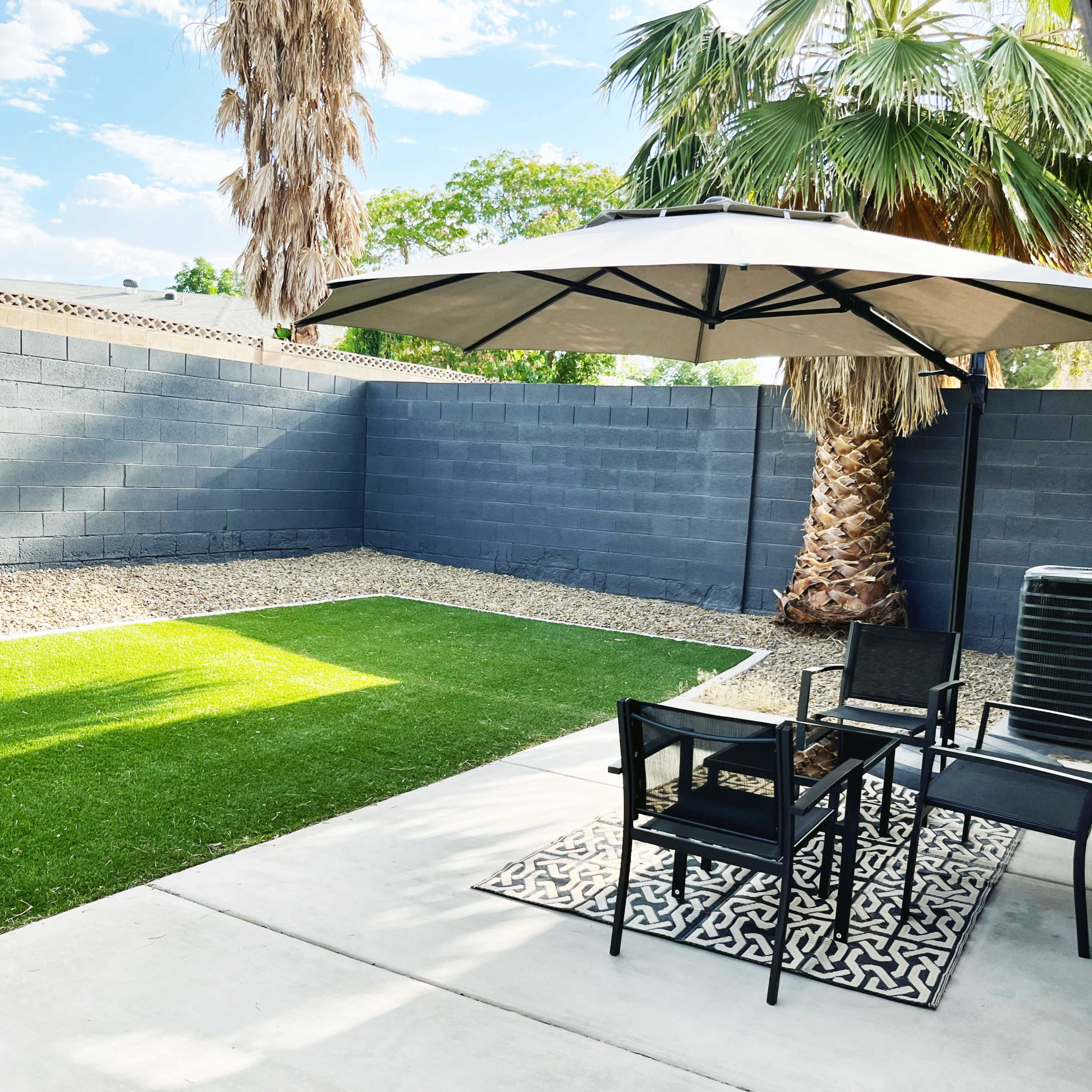 A patio area features a black umbrella over a table and chairs, alongside a landscaped yard with synthetic grass and a palm tree.