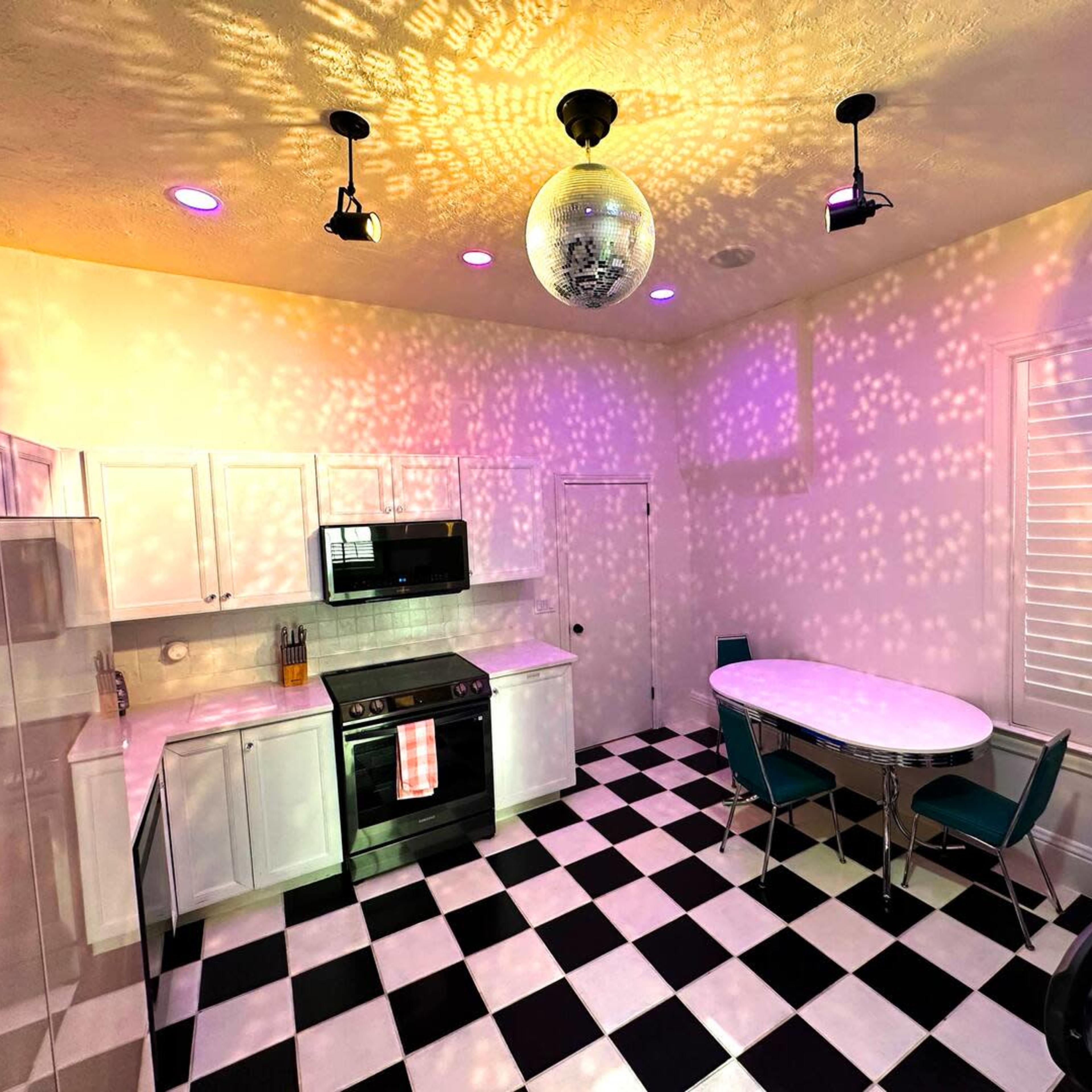 A modern kitchen with black and white checkered flooring, white cabinets, and colorful light reflections on the walls from a disco ball.