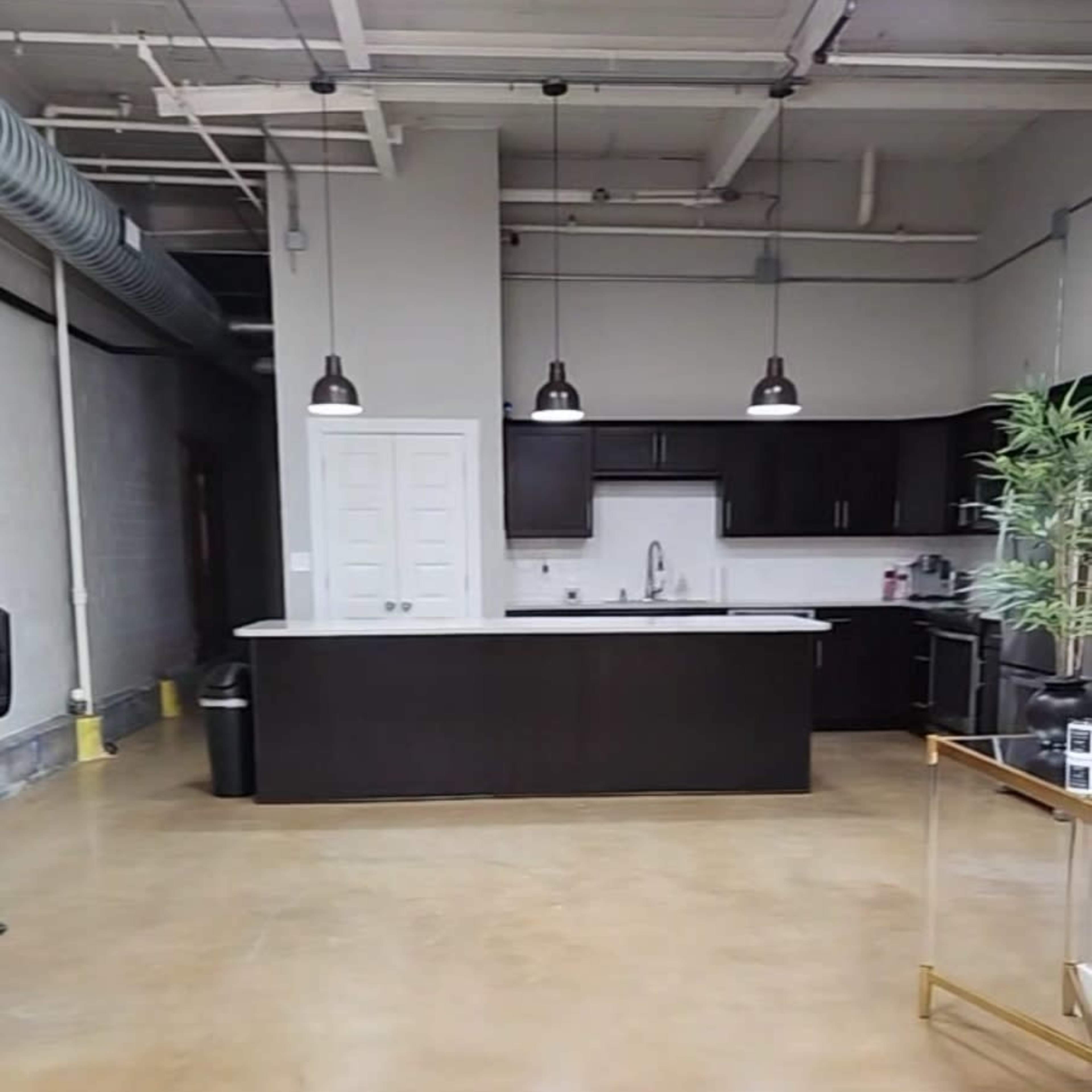 A modern kitchen area with a dark cabinet setup, a white countertop, and industrial-style lighting in an open office space.