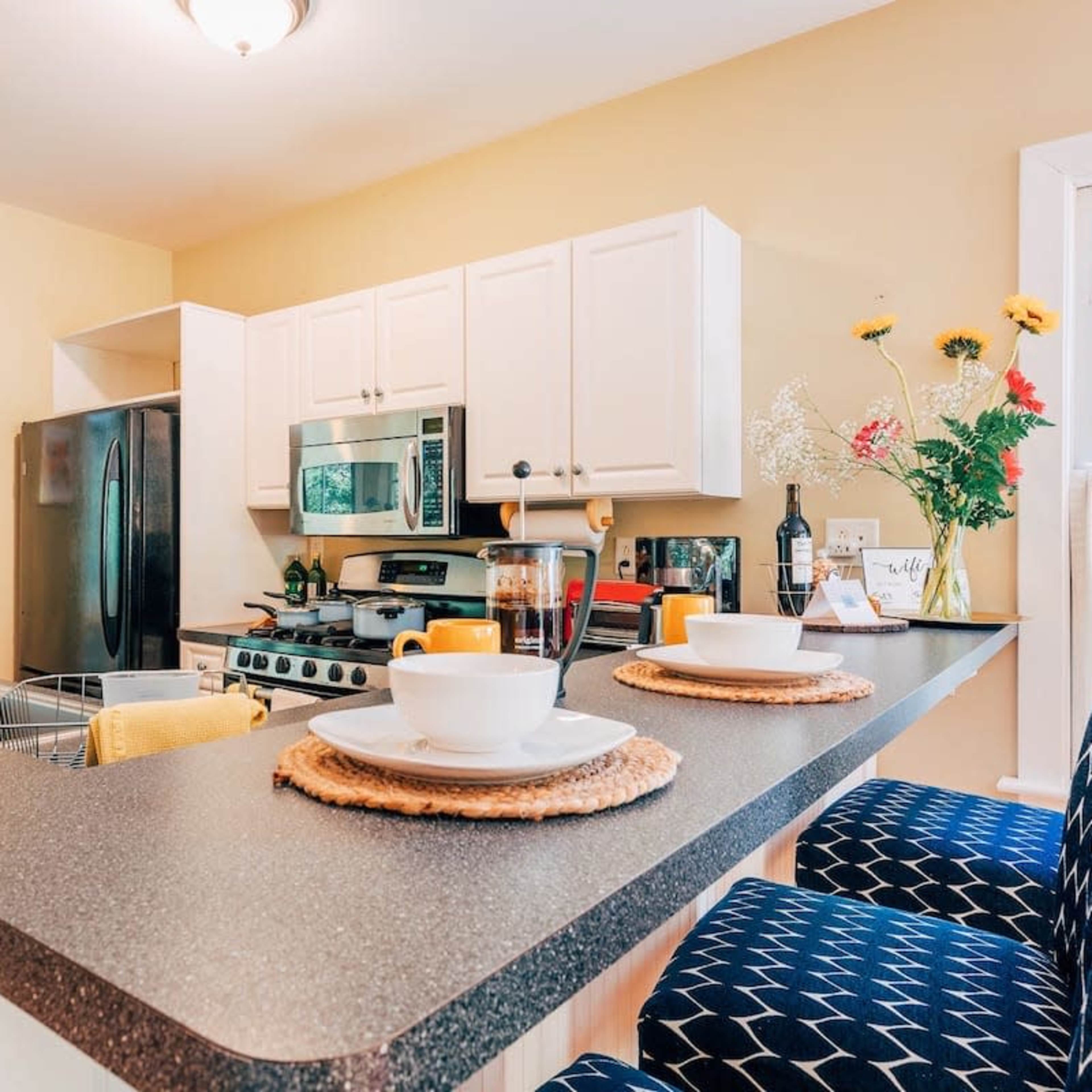 A modern kitchen layout features a countertop with two place settings, a stainless steel stove, and white cabinetry adorned with a vase of flowers.