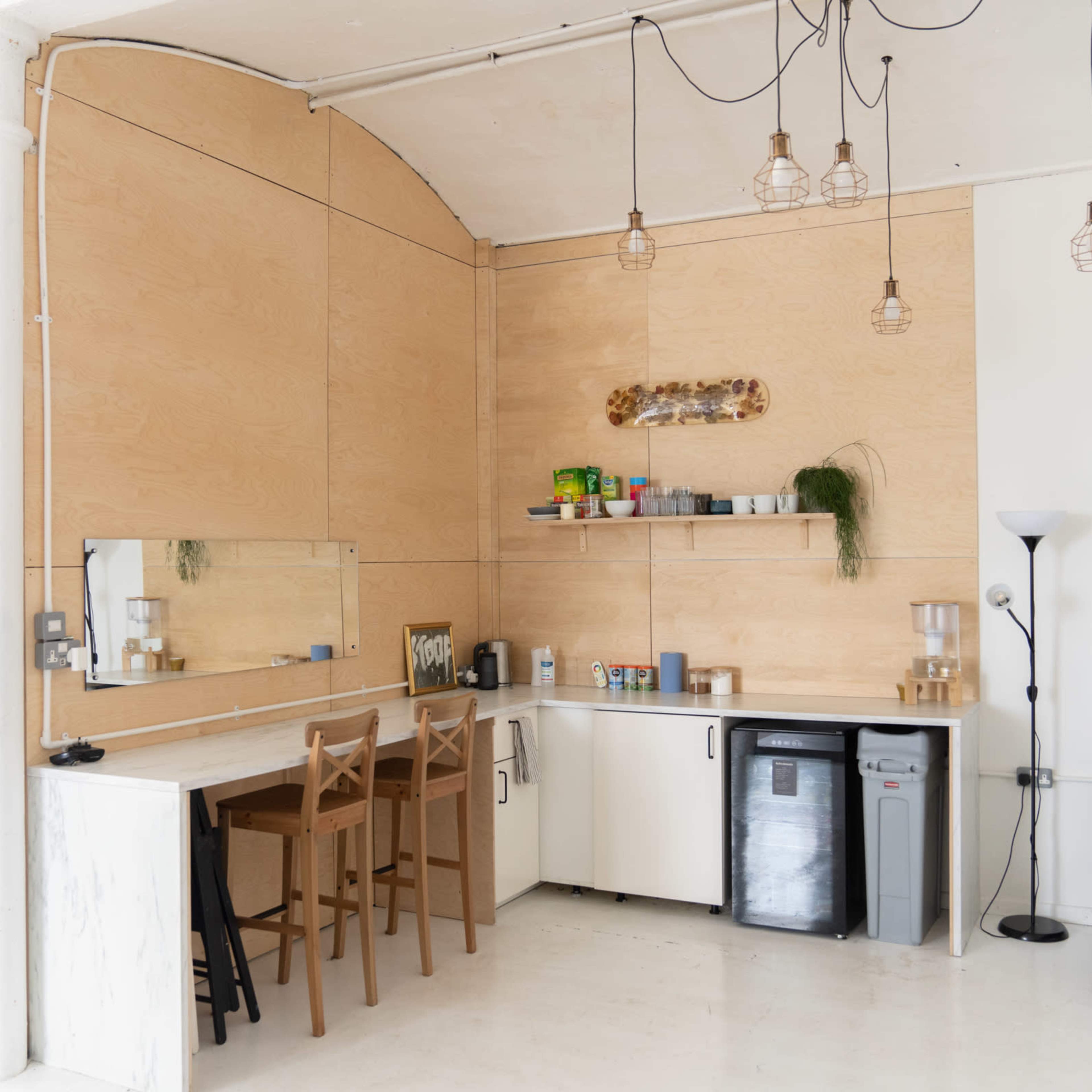A minimalist kitchen area with wooden paneling, a countertop, three bar stools, and a shelf holding plants and kitchen items.