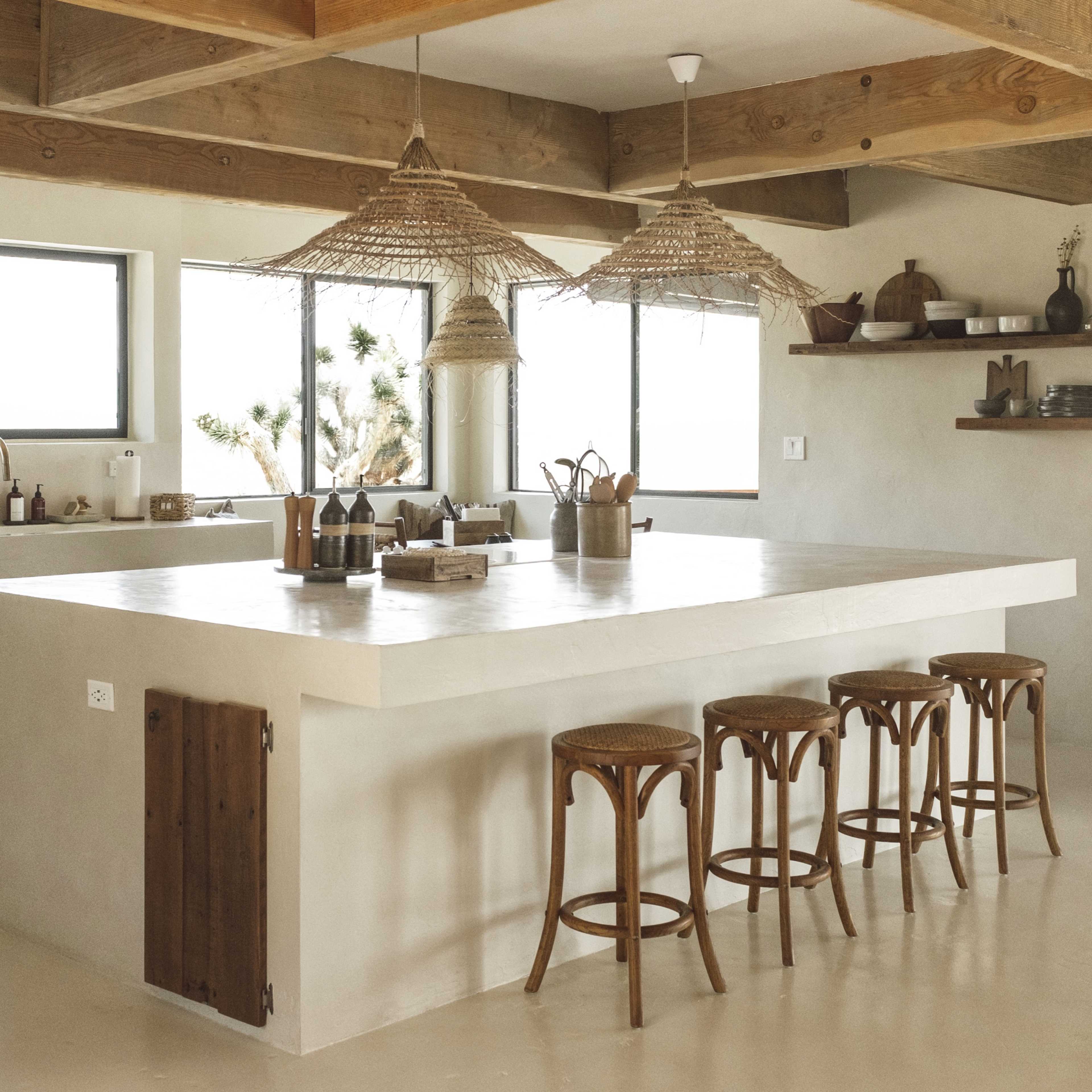 The kitchen features a large island with three wooden stools, under woven pendant lights, surrounded by minimalist decor and large windows.