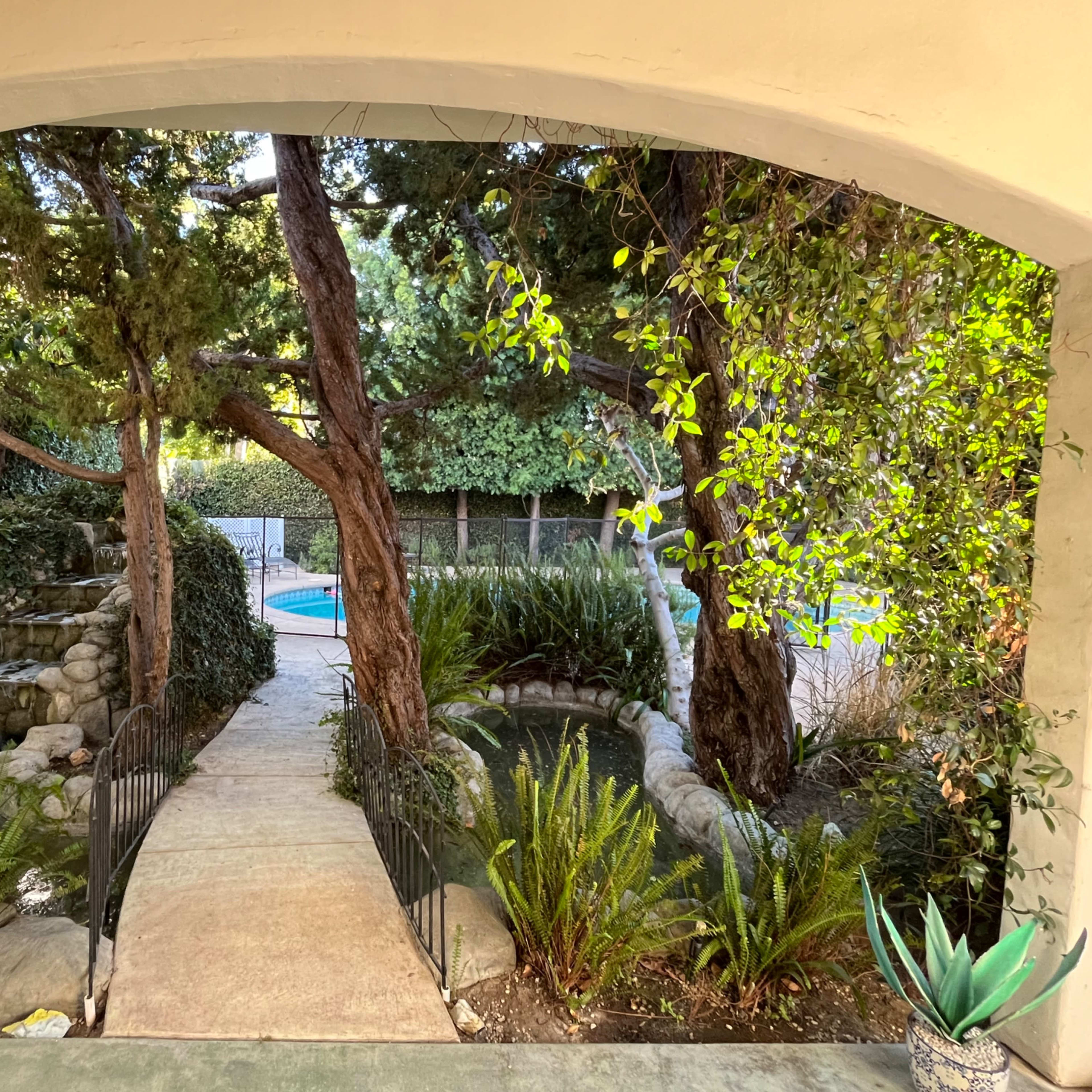 A stone pathway leads through lush greenery to a pool surrounded by trees and shrubs.