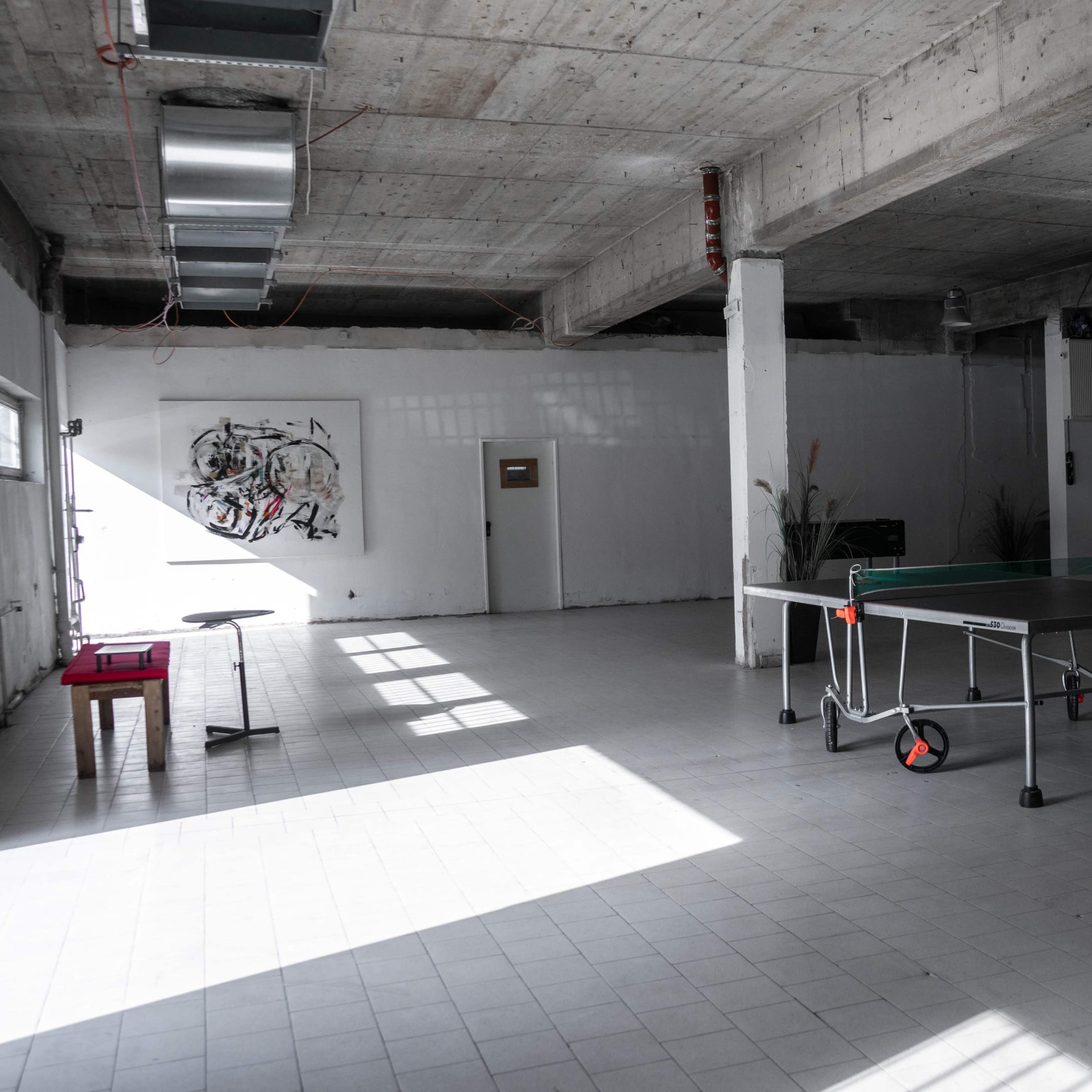 An empty industrial-style room features a ping pong table and a small table near a window, with concrete walls and flooring.