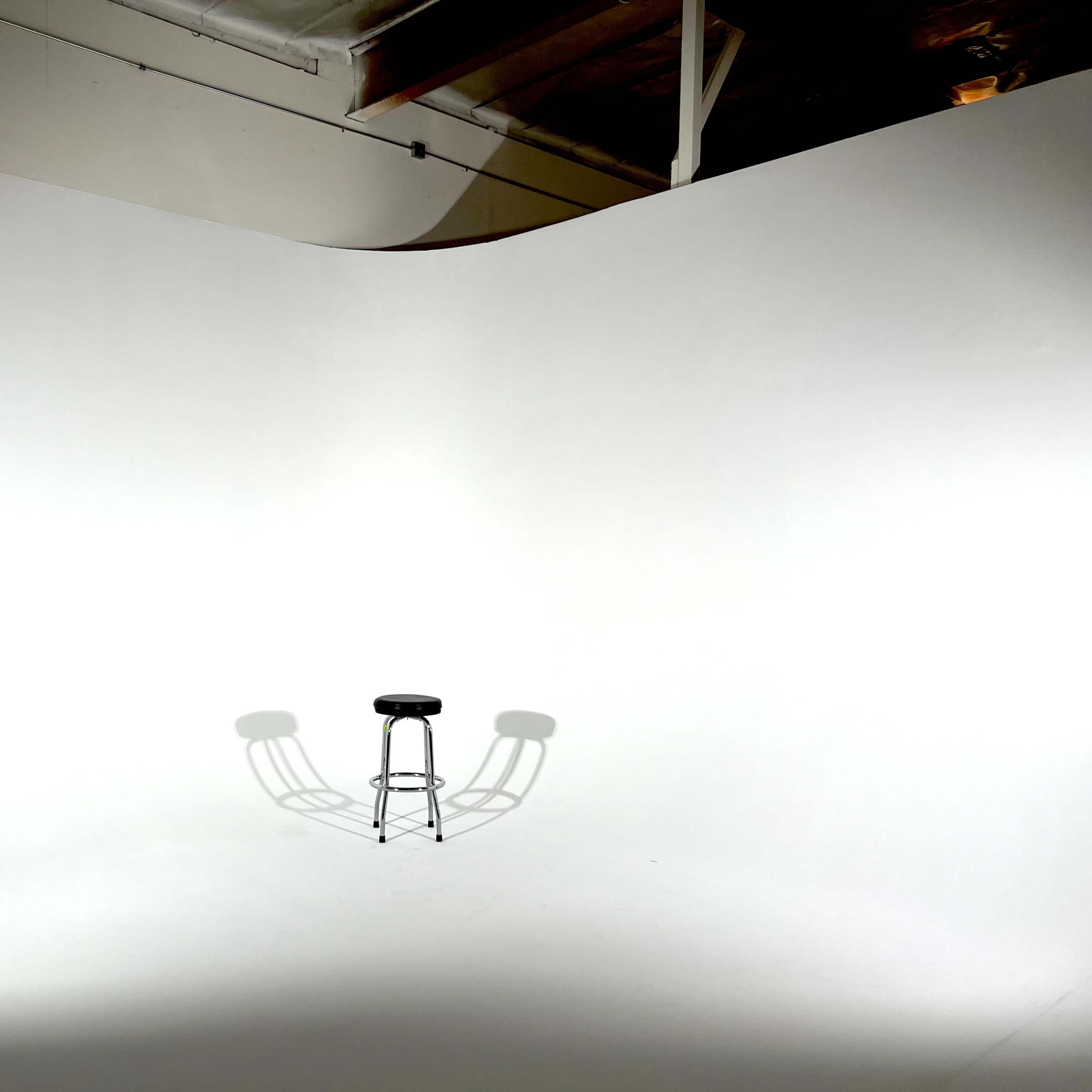 A black stool casts a shadow on a plain white backdrop in an empty studio space.
