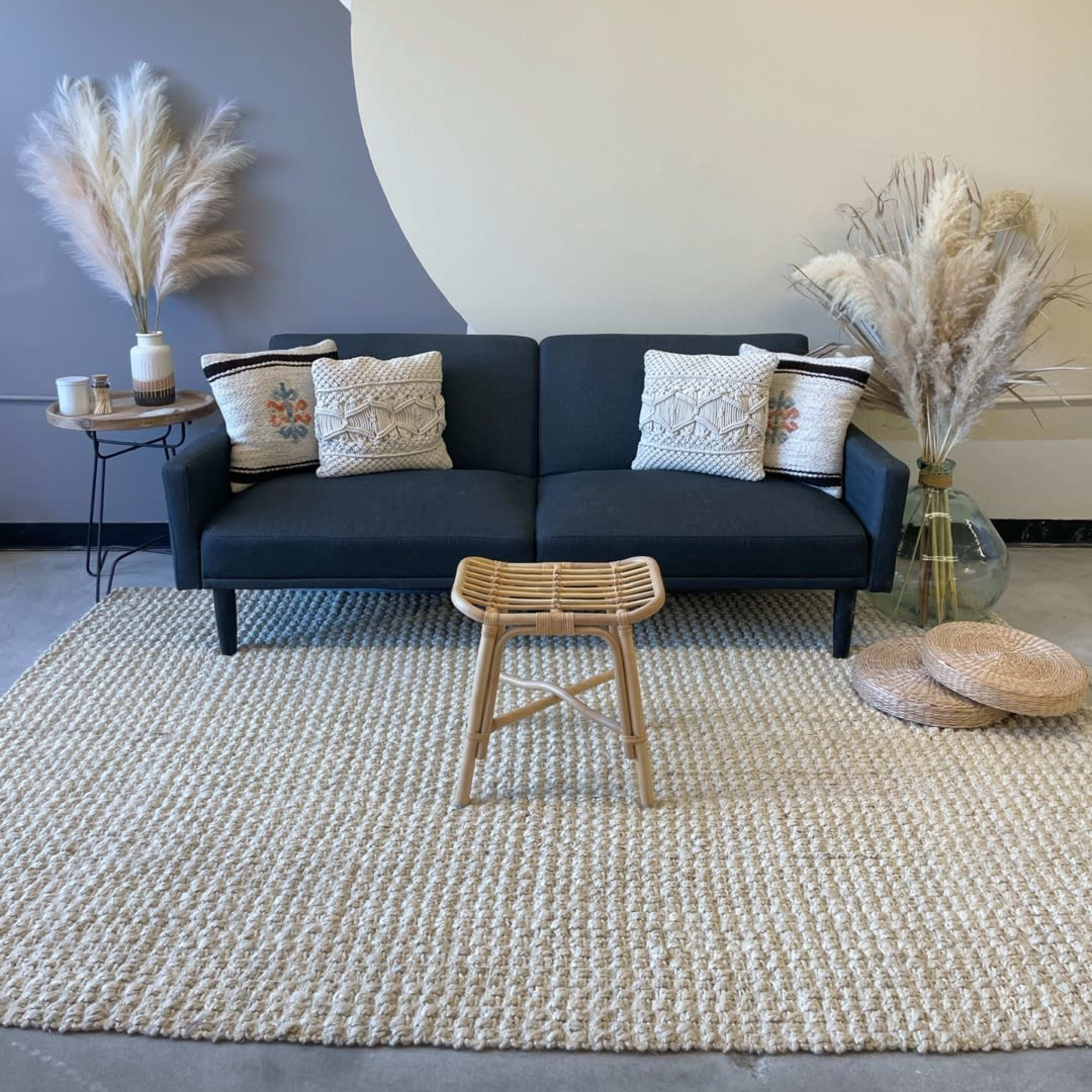 A dark sofa with decorative pillows is positioned on a textured rug, accompanied by a small stool and tall decorative grasses in vases.
