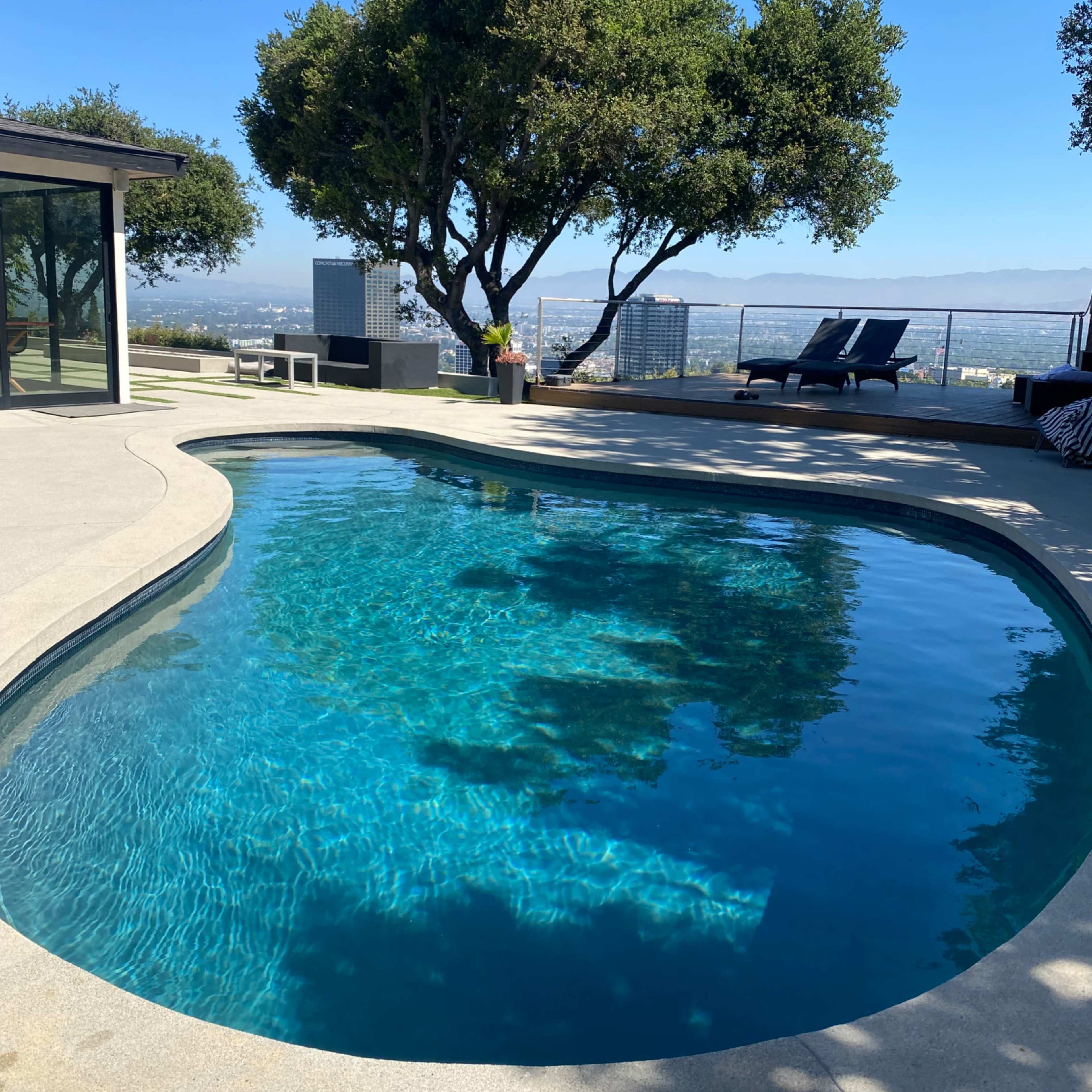 A curved swimming pool surrounded by trees and lounge chairs offers a view of a cityscape under a clear blue sky.