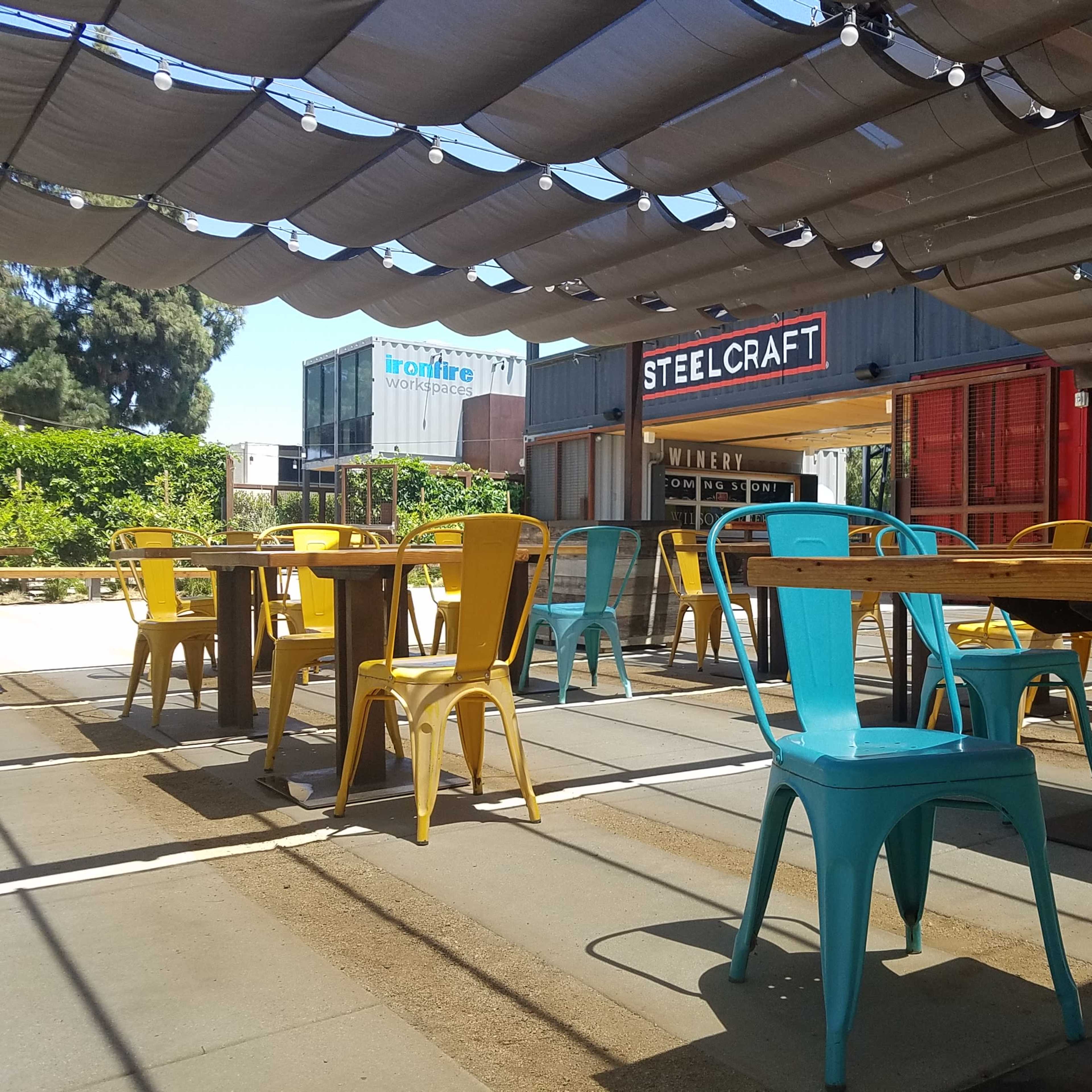 An outdoor seating area at a venue with colorful metal chairs and tables under a shaded canopy.