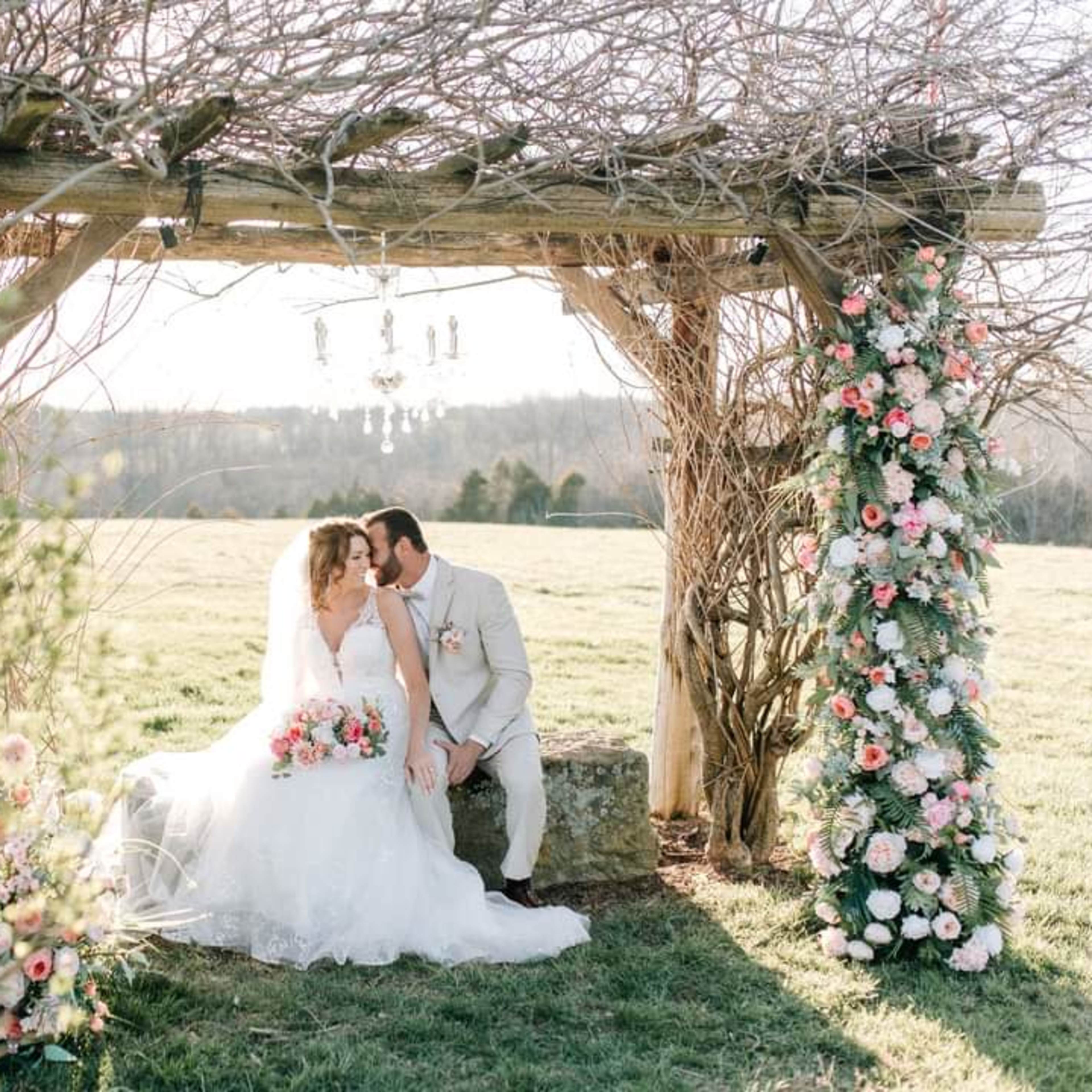 A couple sits under a decorated arbor, surrounded by flowers, while sharing a moment of intimacy in a grassy outdoor setting.