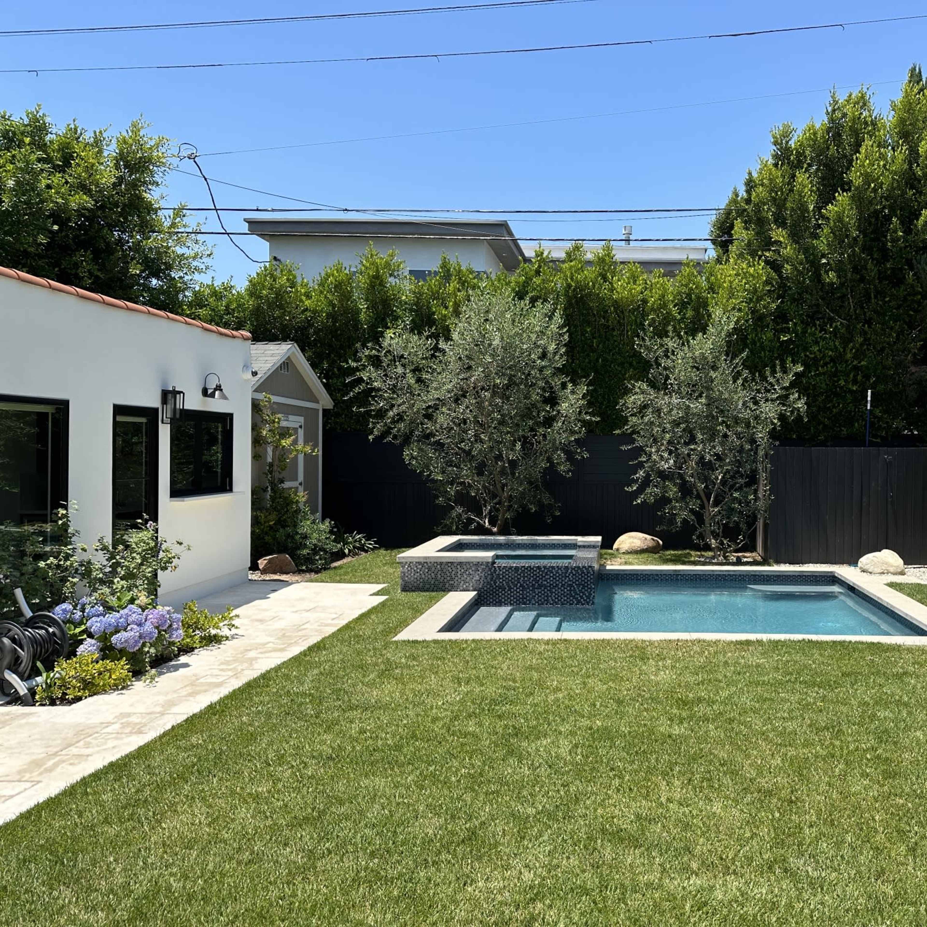 A landscaped backyard featuring a rectangular swimming pool, a small hot tub, and neatly trimmed grass surrounded by trees and shrubs.