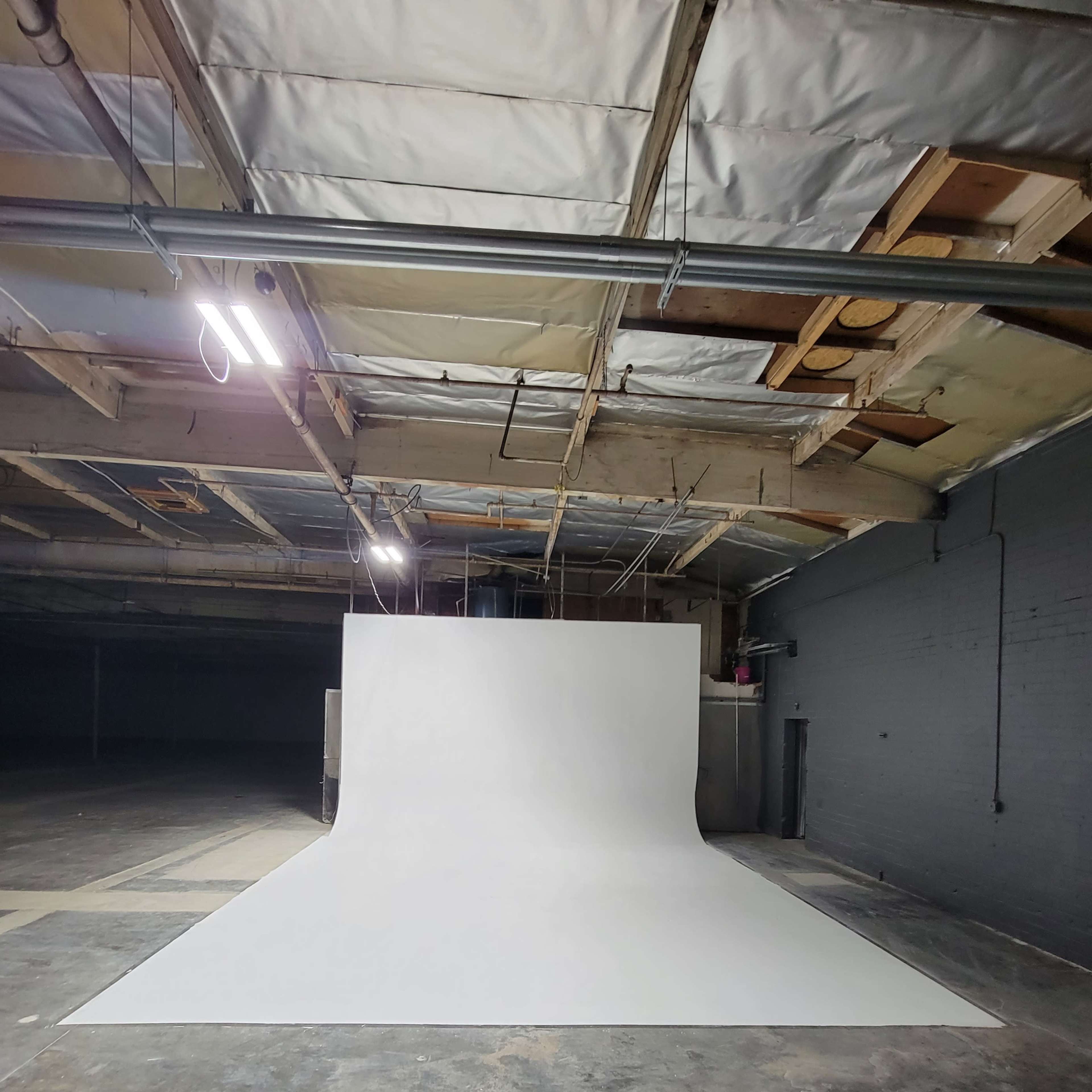 A large, white seamless backdrop is set up in a dimly lit industrial space with exposed beams and concrete flooring.