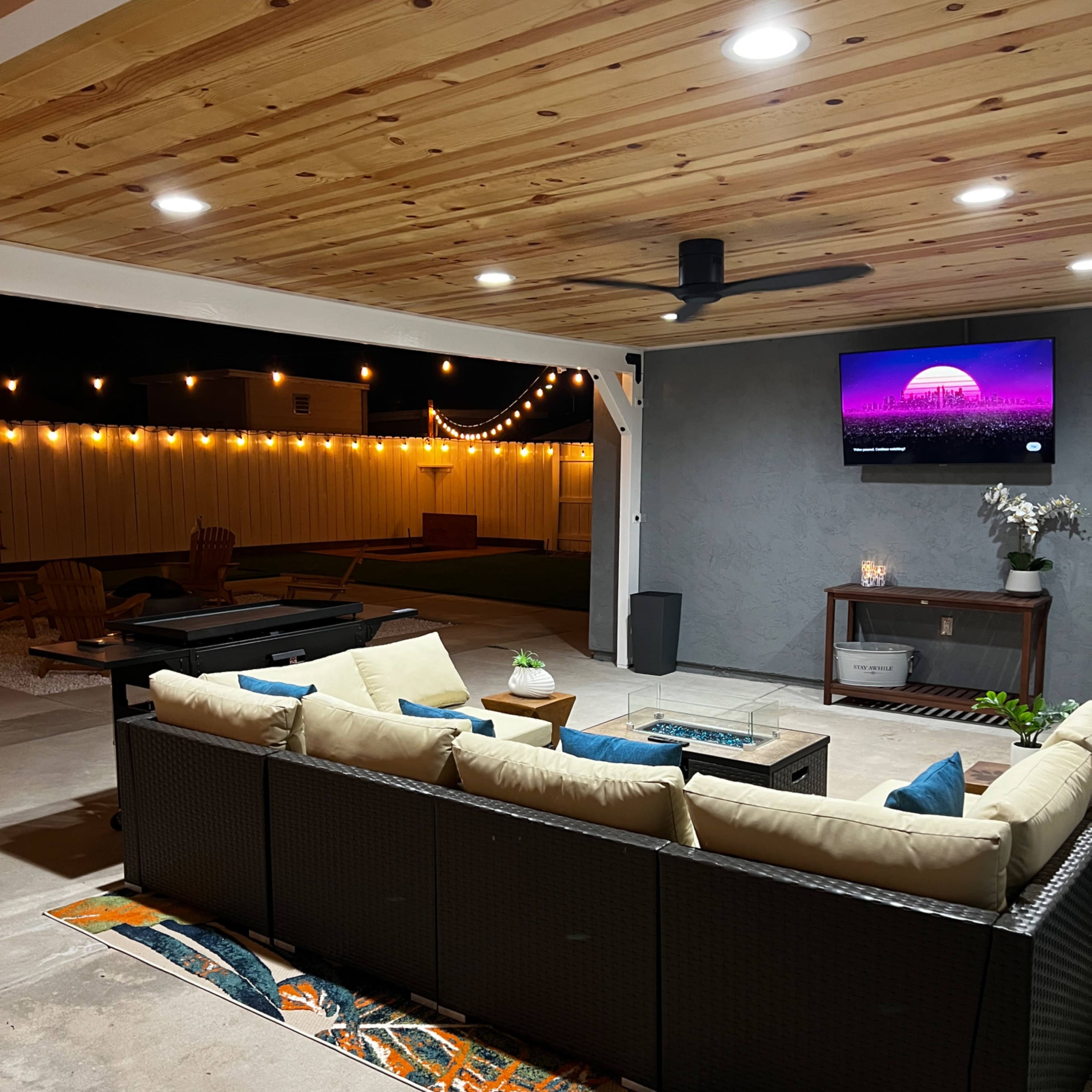 A patio area features a sectional sofa with light-colored cushions, a coffee table, and a wall-mounted TV, illuminated by string lights overhead.