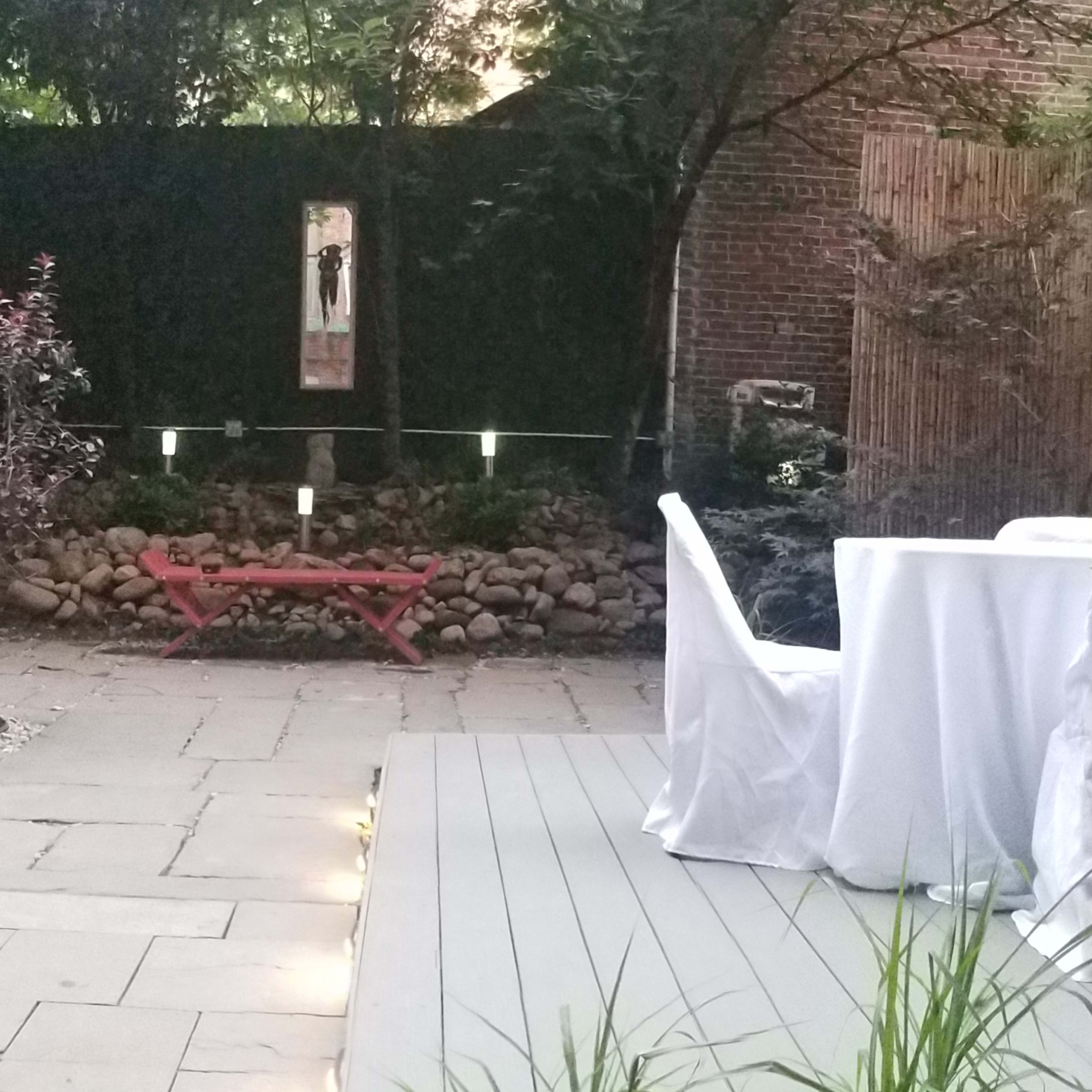 A small, landscaped outdoor space with stone pathways, a seating area featuring a table covered with a white cloth, and various plants and rocks surrounding the area.