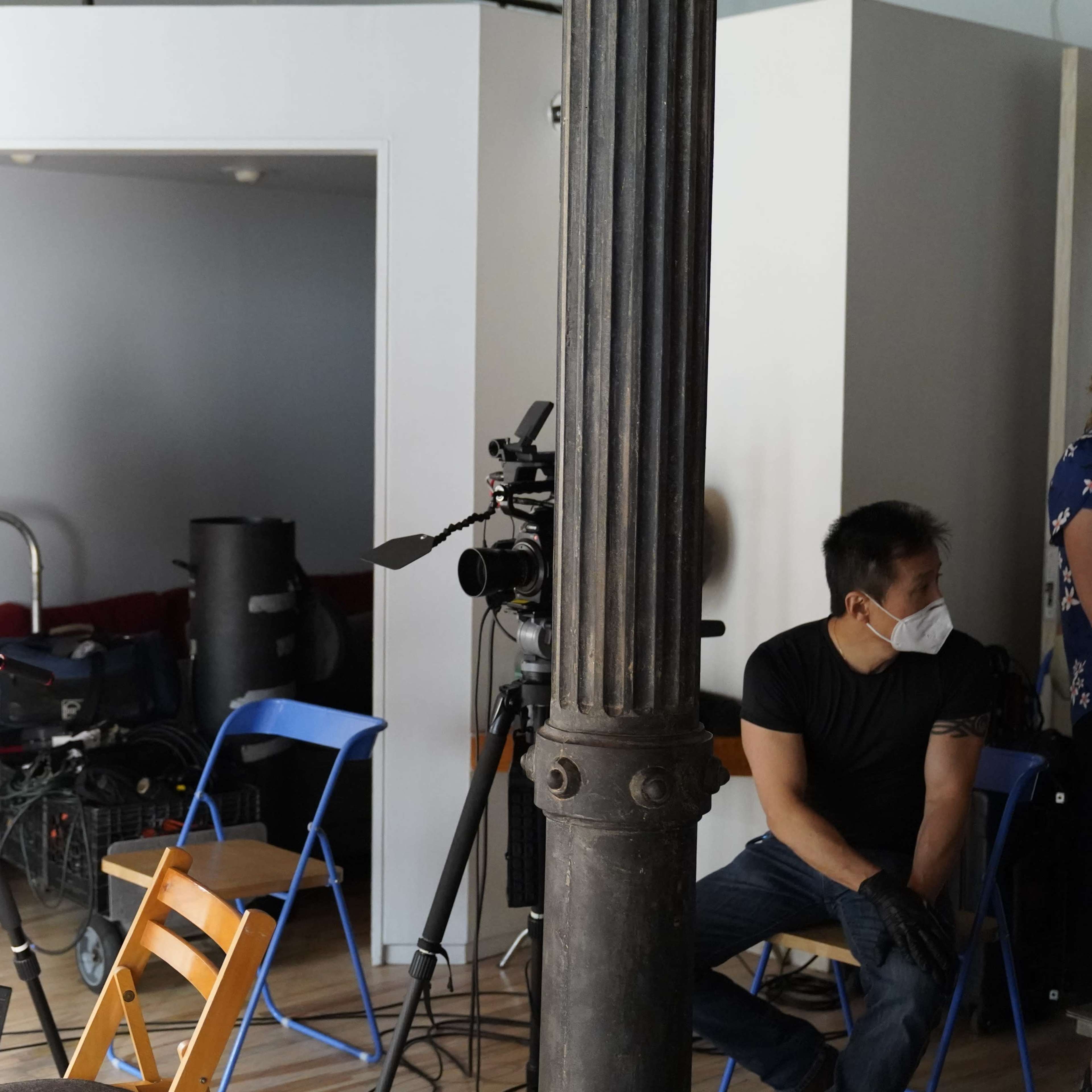 A small film set with two cameras on tripods, a man wearing a mask sitting on a blue chair, and a column in the foreground.