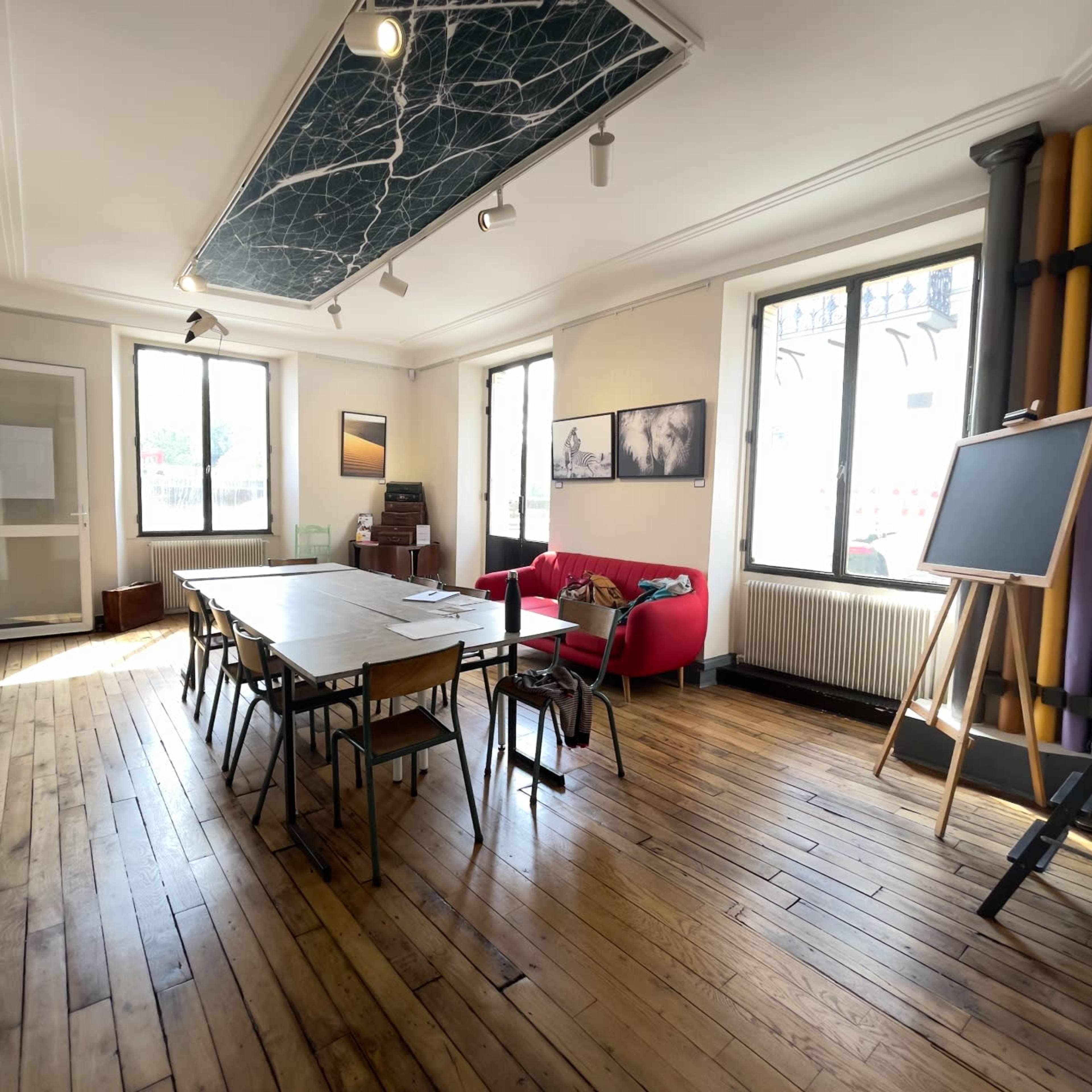A bright meeting room with wooden floors, a long table surrounded by chairs, a red couch, large windows, and a chalkboard.