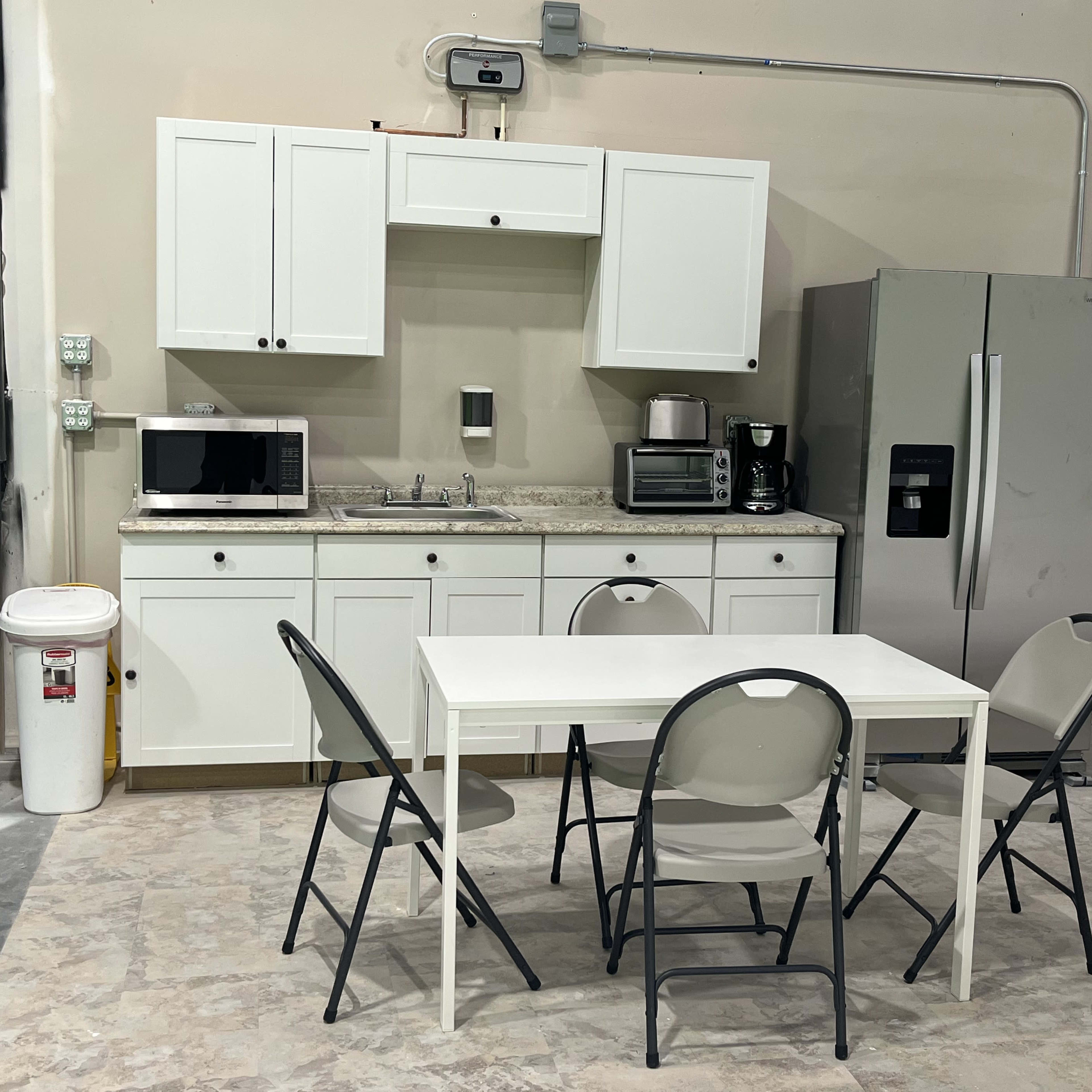 A simple kitchen area features white cabinets, a sink, a microwave, a coffee maker, a stainless steel refrigerator, and a table surrounded by four folding chairs.
