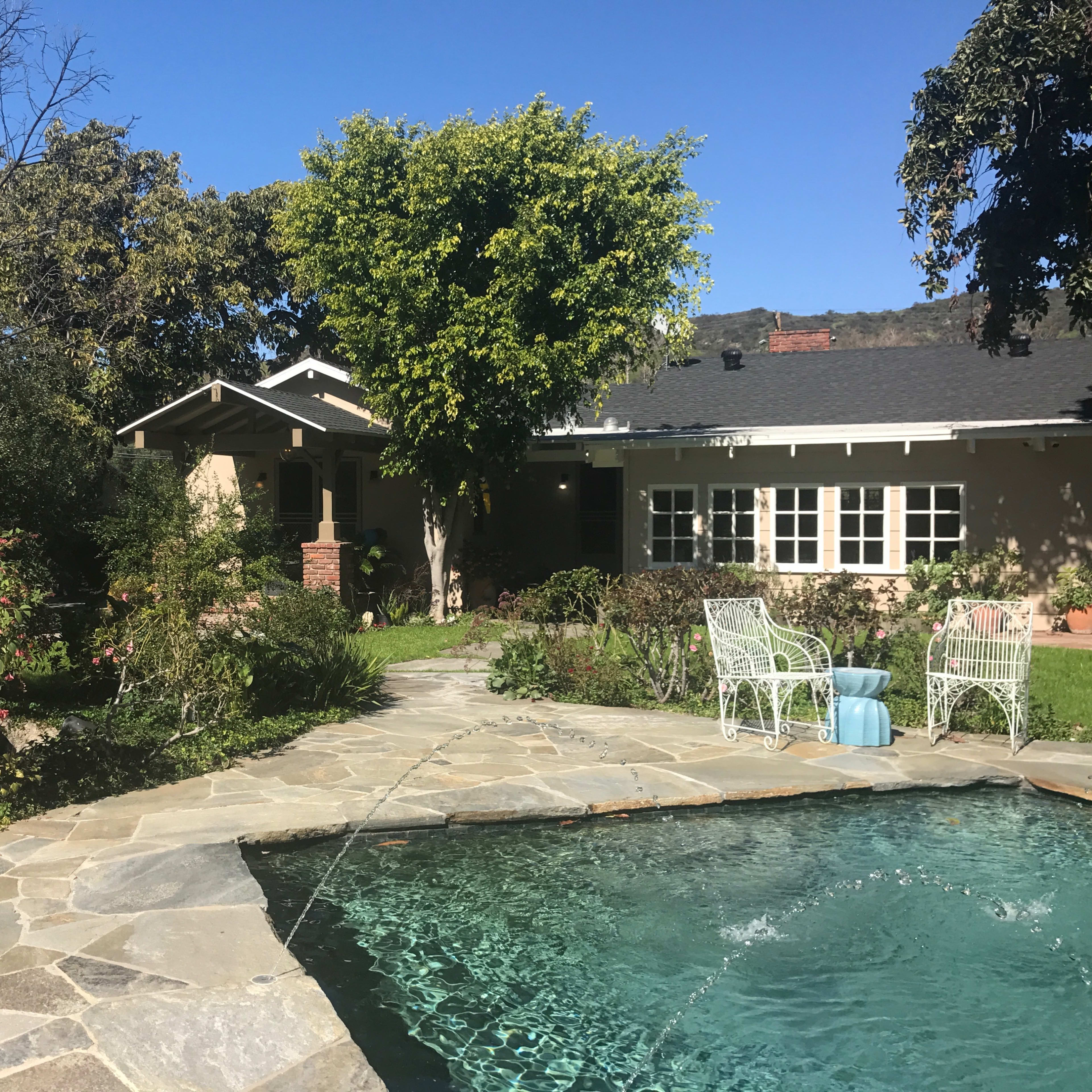 A landscaped backyard with a stone patio, a swimming pool, and a house partially obscured by trees and shrubs.