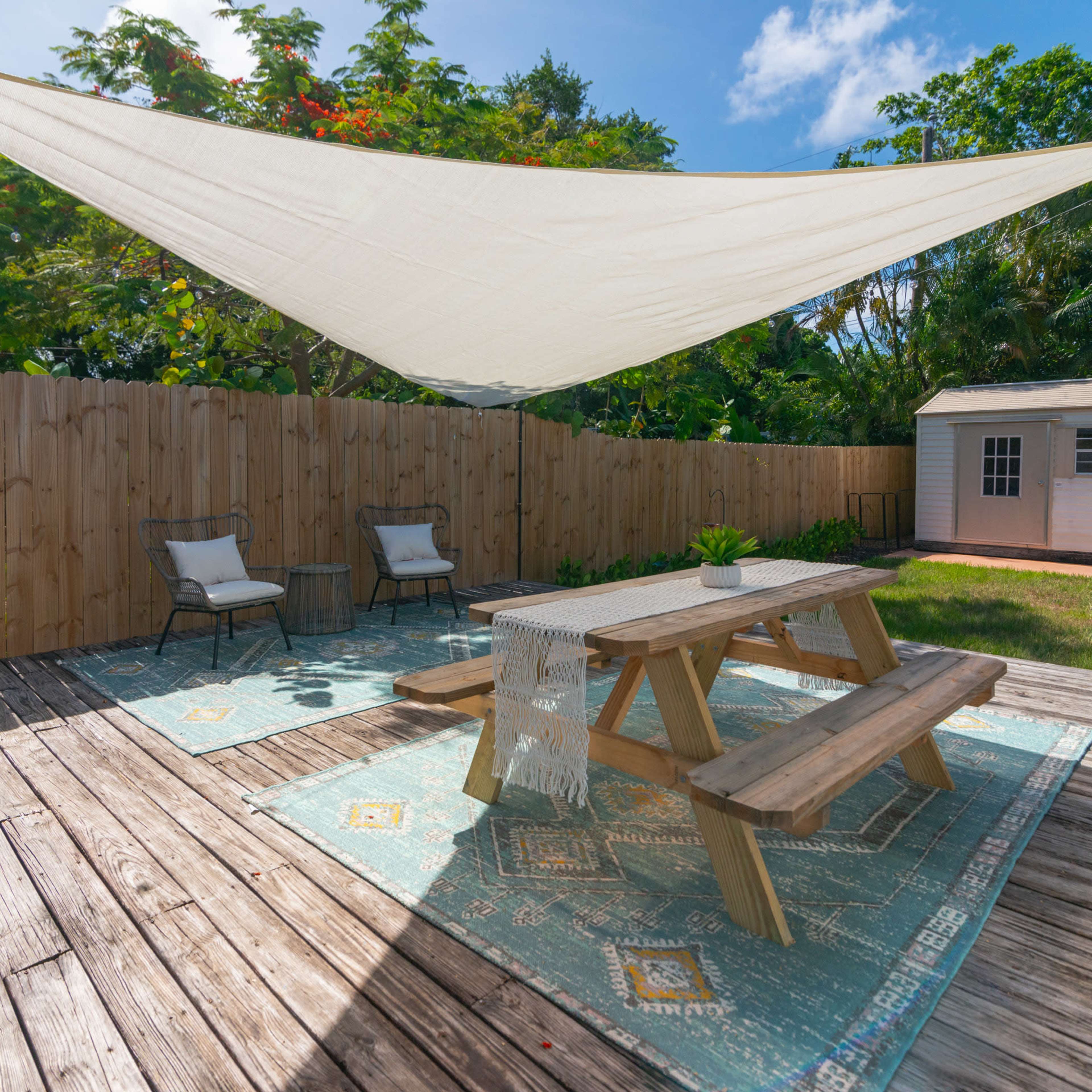 A shaded outdoor patio features a wooden table and chairs on a rug, surrounded by a wooden fence and greenery.