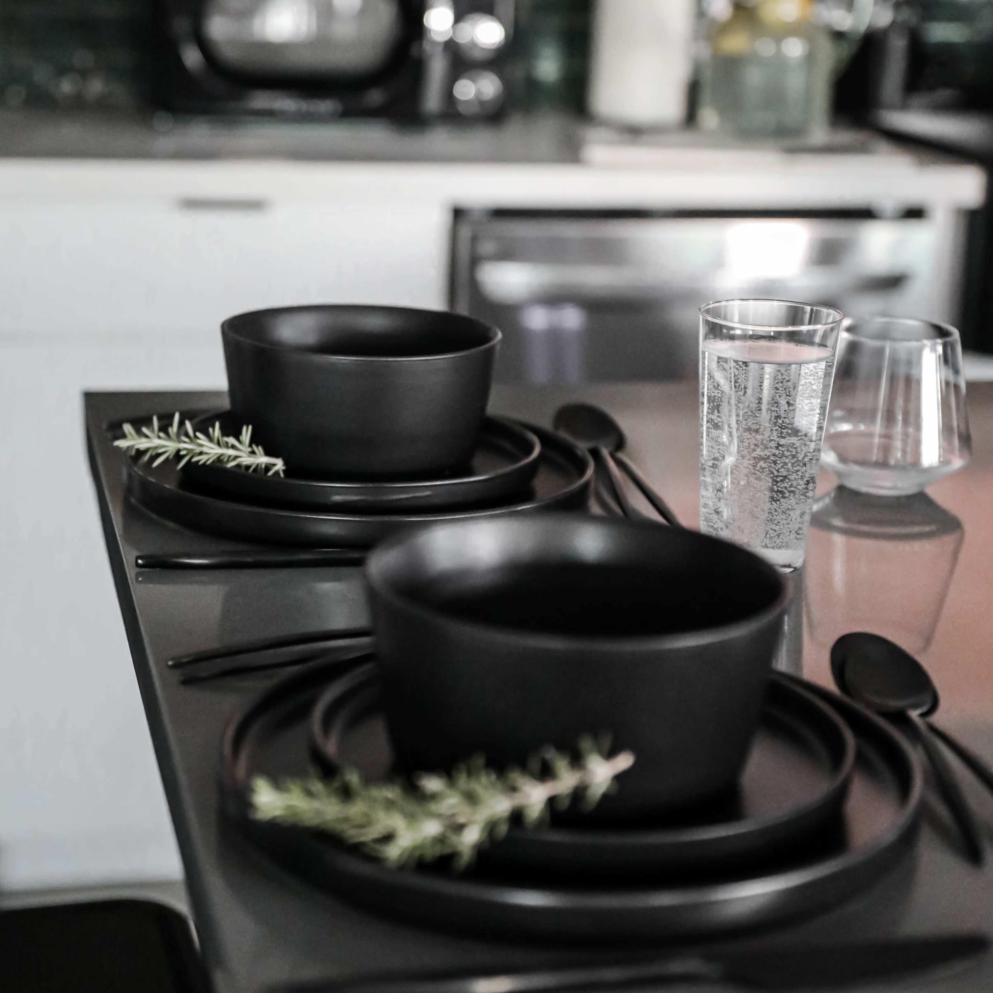 A dark kitchen countertop is set with two black bowls, black plates, and utensils, garnished with sprigs of rosemary, alongside a glass of water and a clear tumbler.