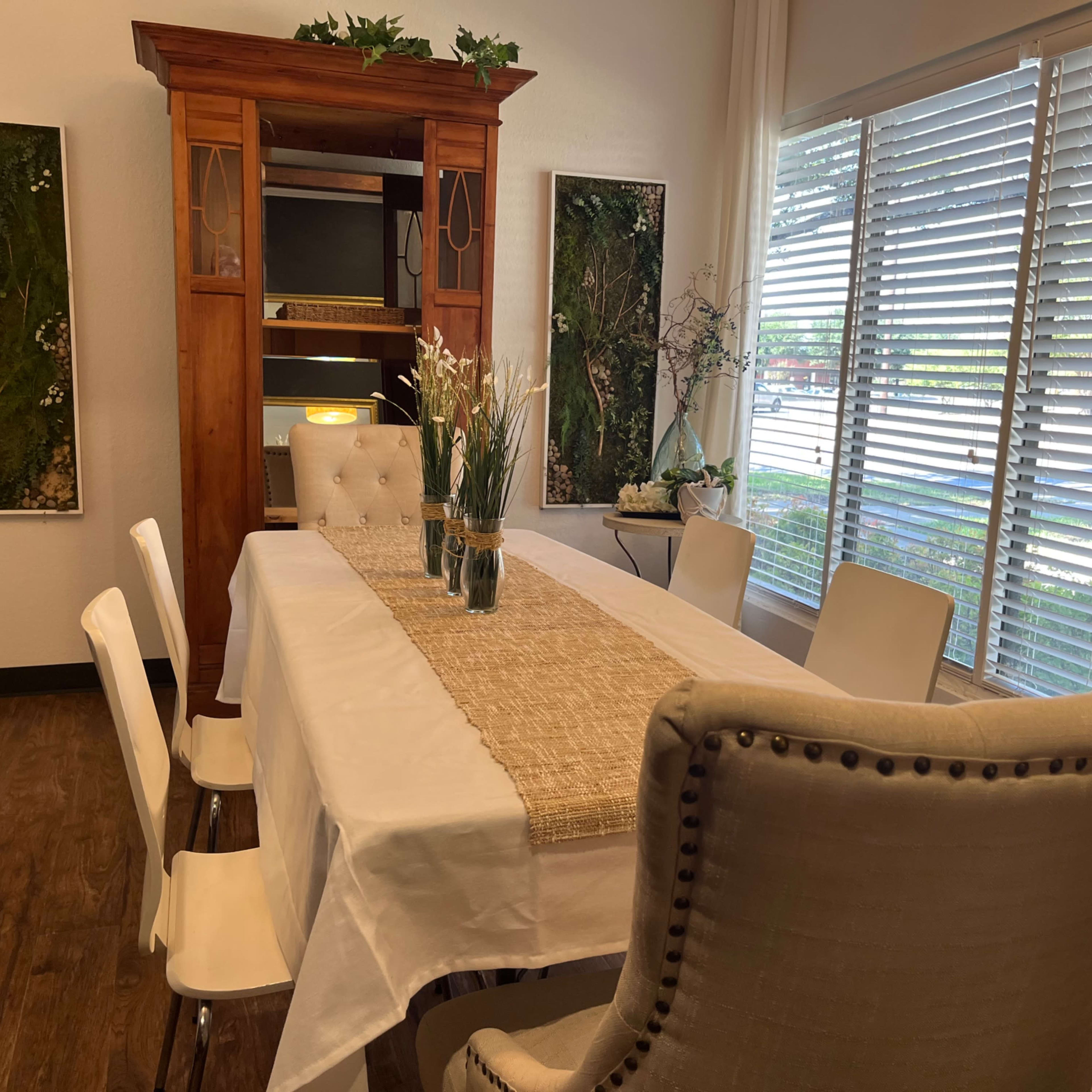 A dining area features a long table set with a table runner, surrounded by white chairs and a tufted armchair, complemented by shelves and framed greenery on the walls.