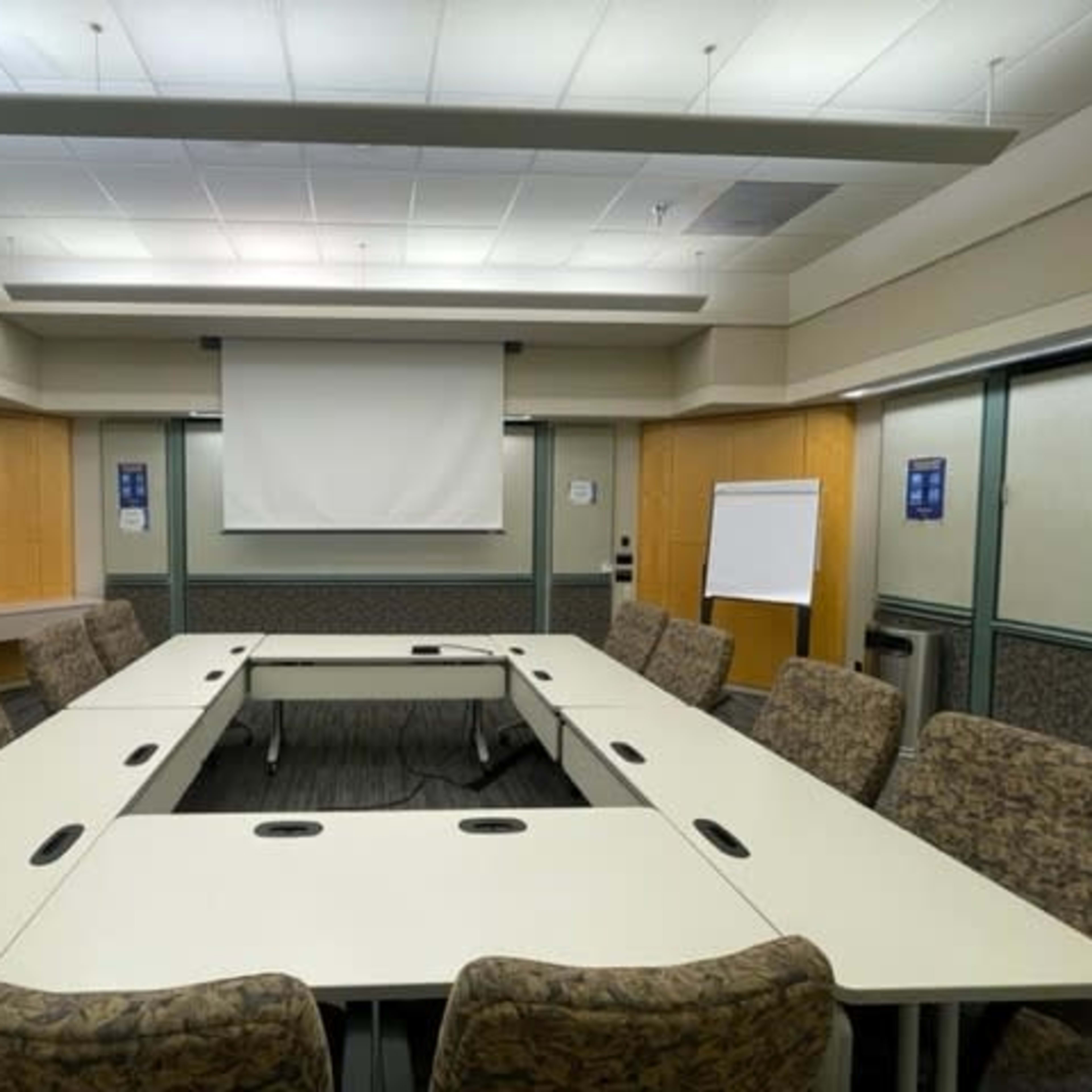A conference room with a large U-shaped table, chairs arranged around it, and a projector screen at the front.