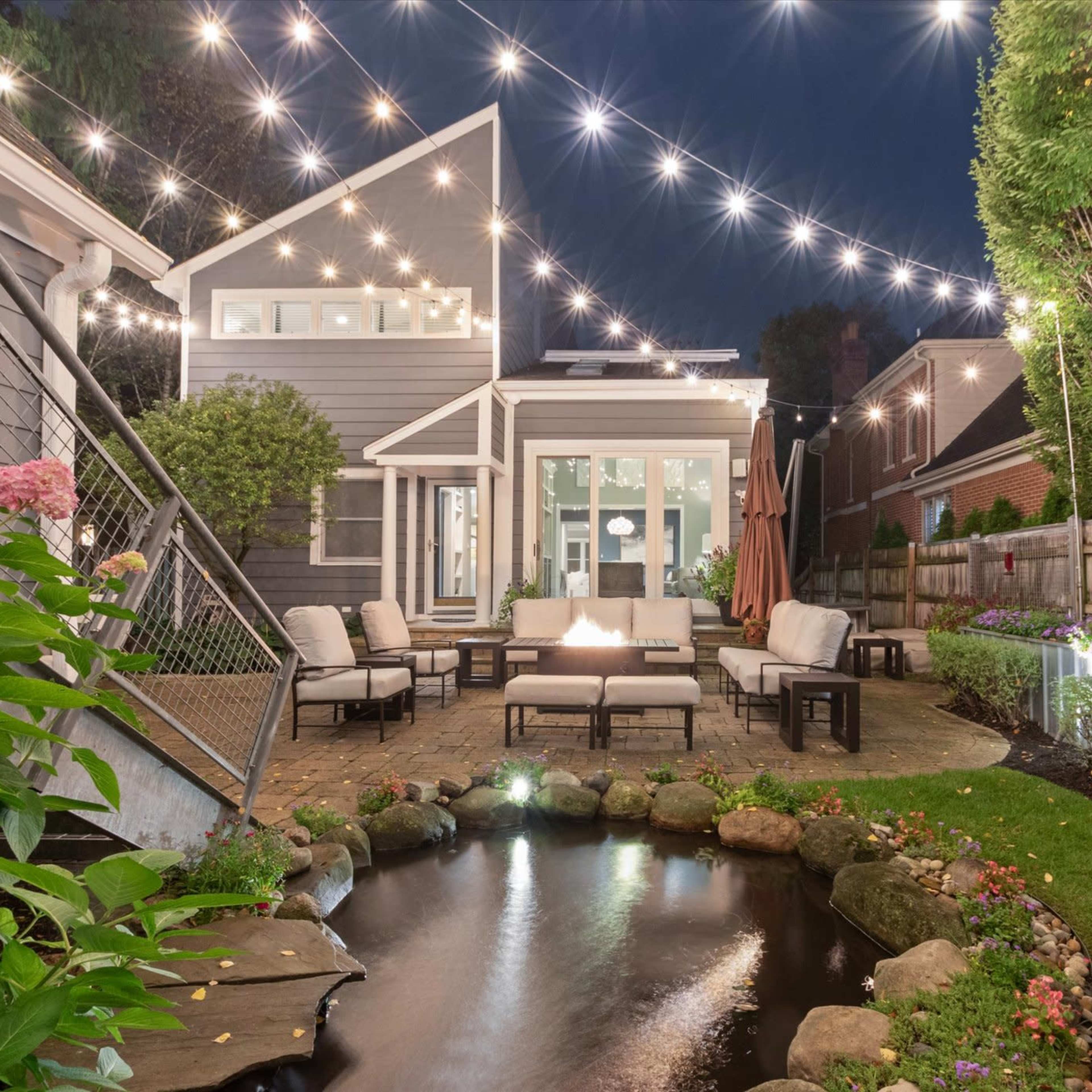 A well-lit backyard features a seating area with a fire pit, surrounded by a pond and neatly arranged landscaping.