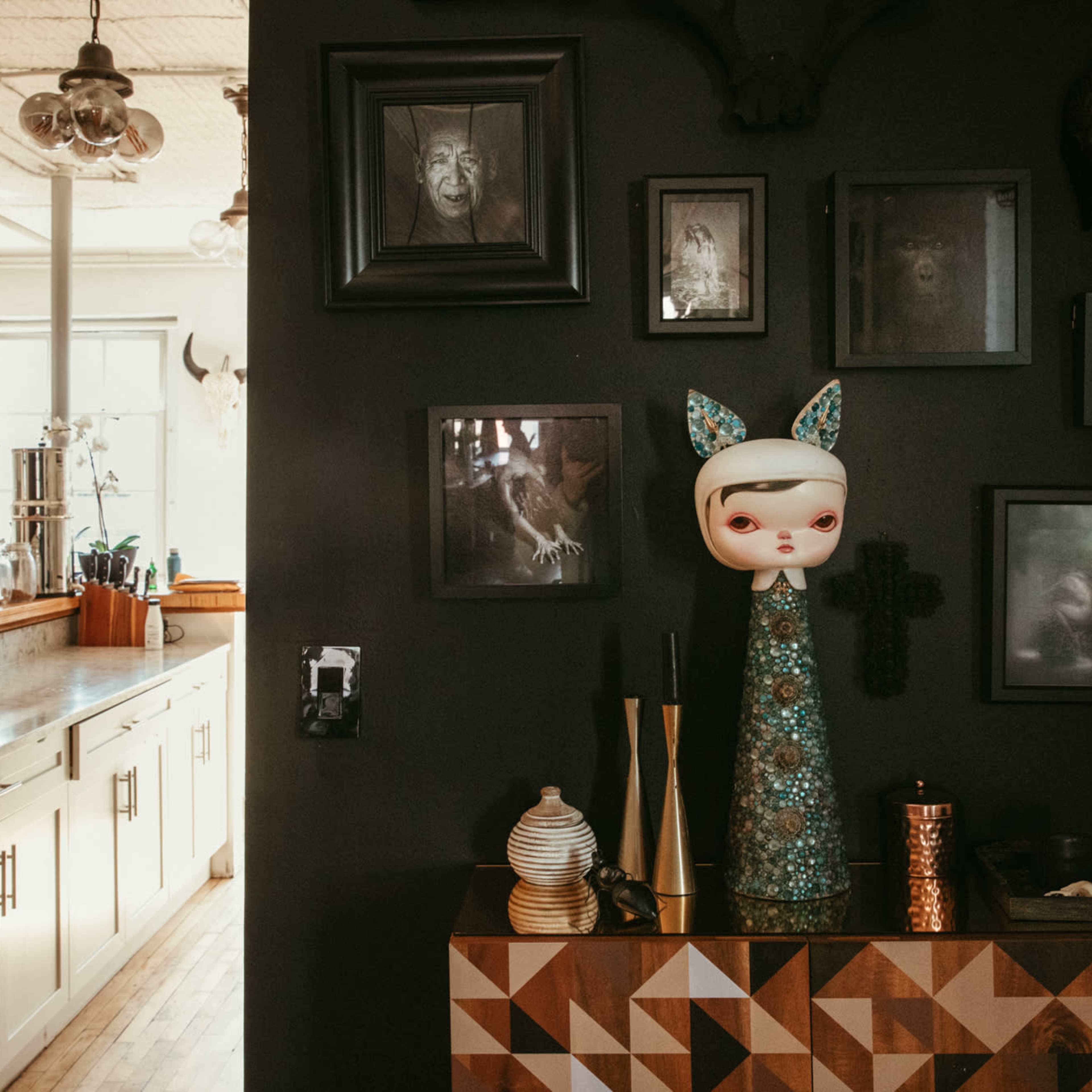 A stylish interior with a dark wall displaying framed art and a decorative figure on a geometric-patterned cabinet, complemented by a bright kitchen area in the background.
