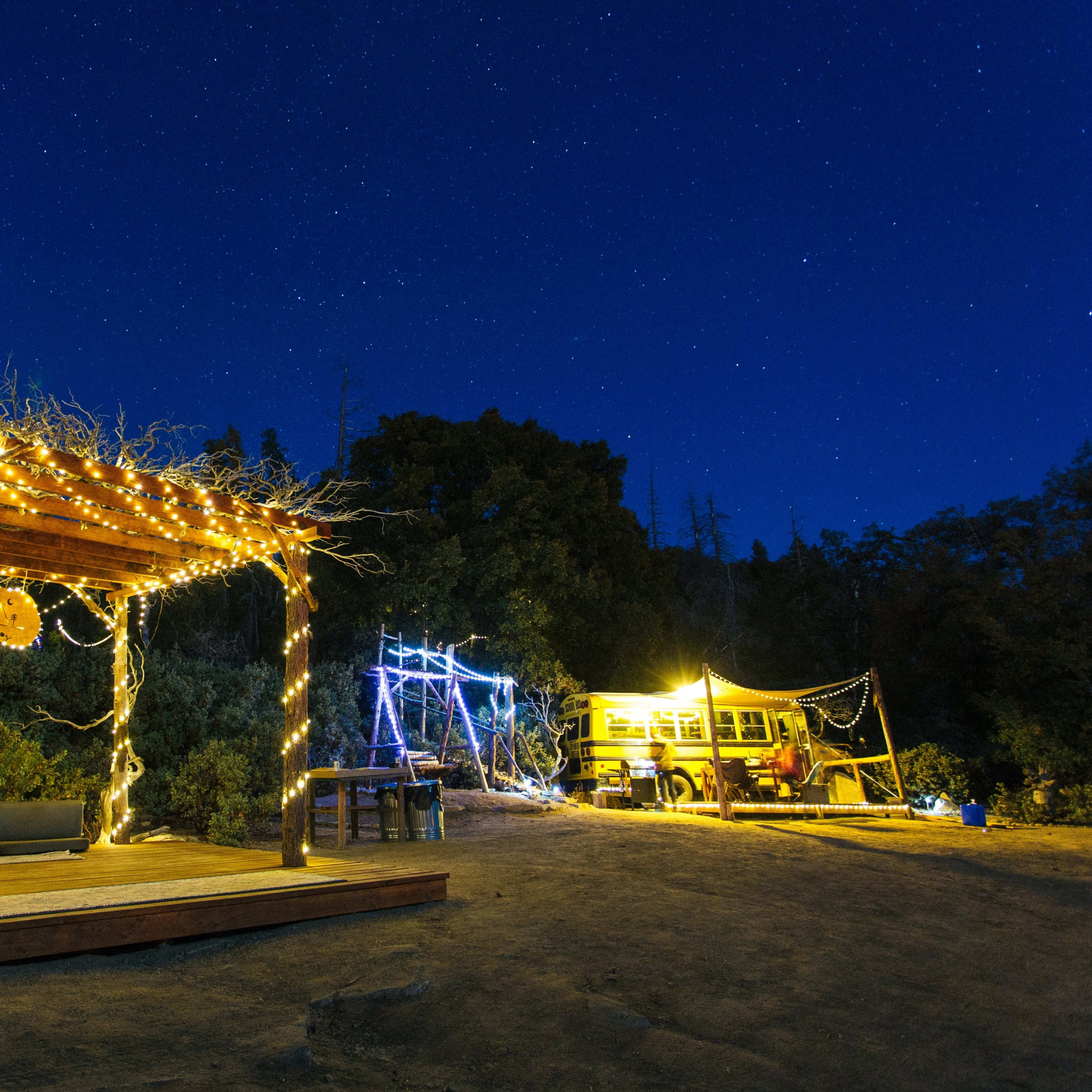 A wooden deck with string lights surrounds a sofa, while a yellow bus is illuminated nearby under a starry night sky.