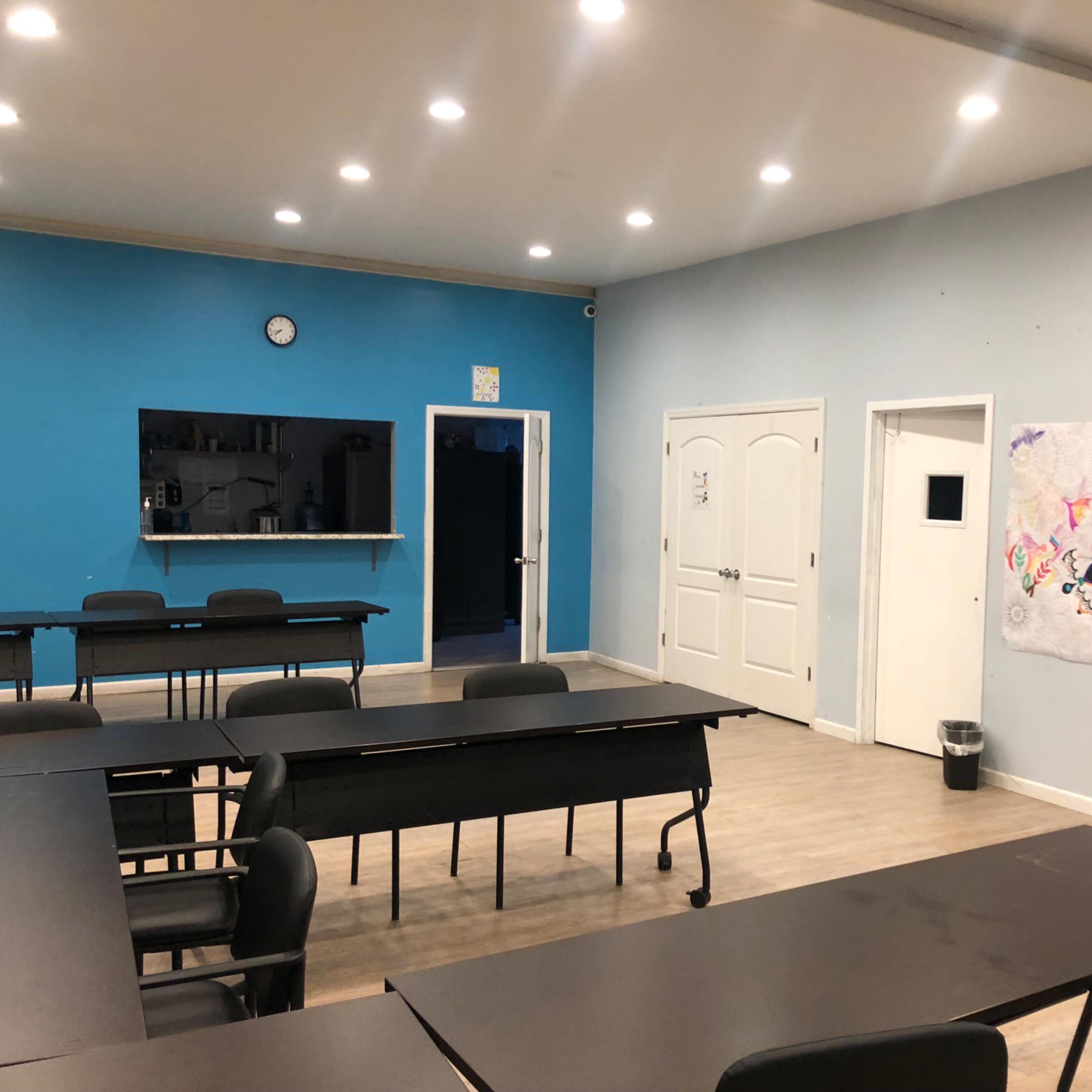 A classroom features several black desks arranged in rows, with blue and light gray walls and a large piece of artwork on one wall.