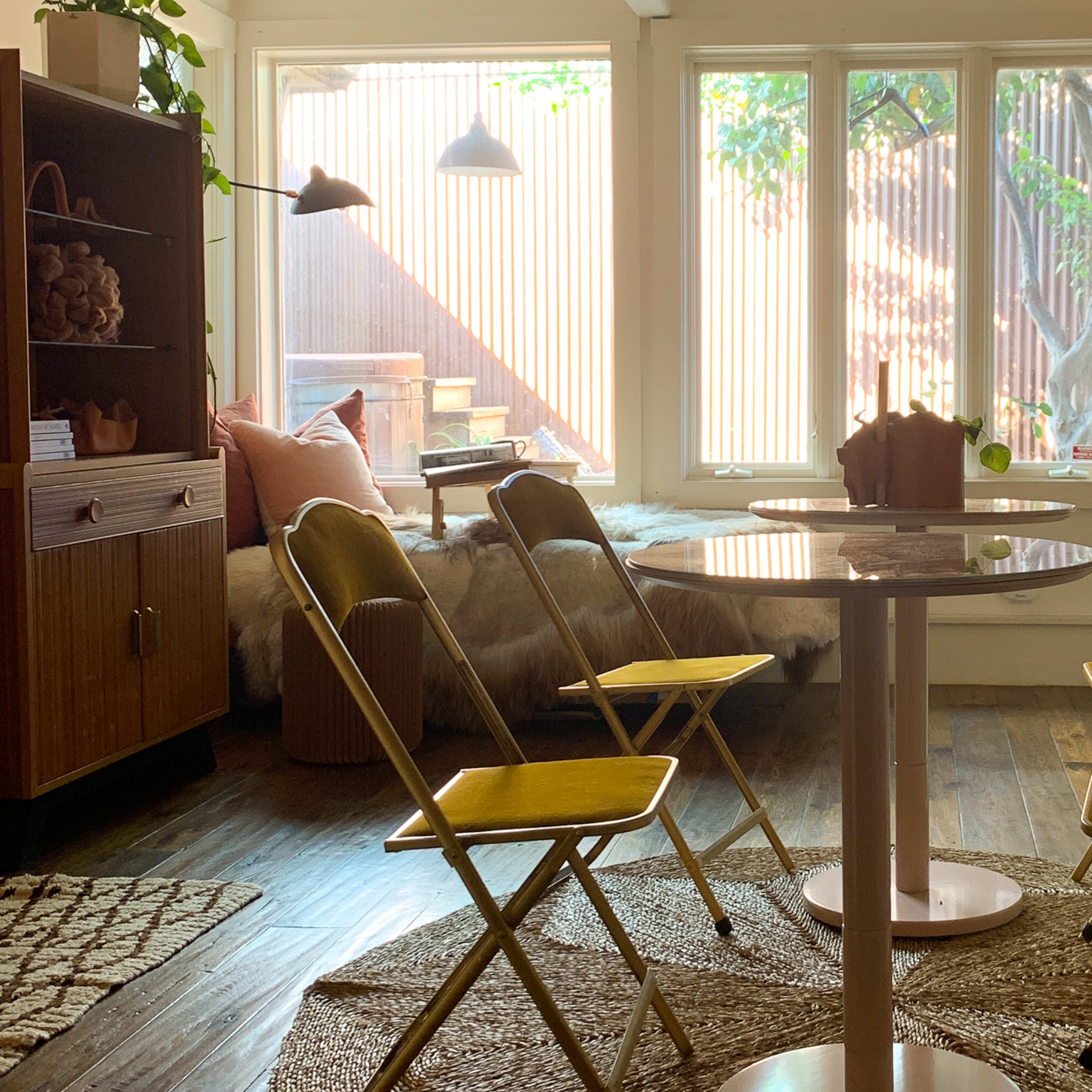 A cozy room with a circular table surrounded by four yellow folding chairs, soft seating, and large windows letting in natural light.